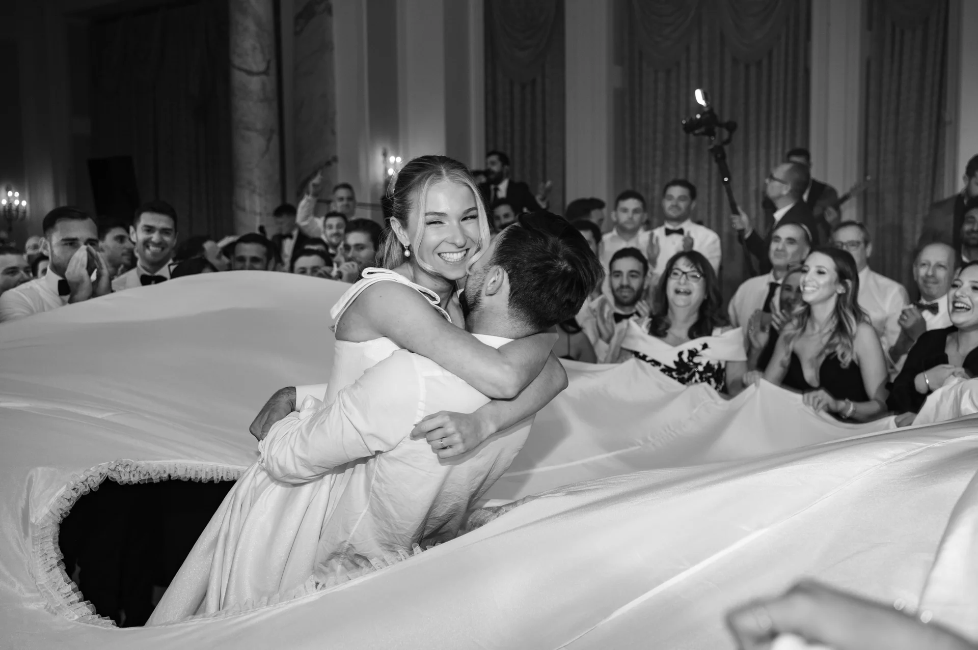 A joyful groom lifts his smiling bride in the air as guests cheer and hold up a large white fabric sheet during a lively wedding reception.