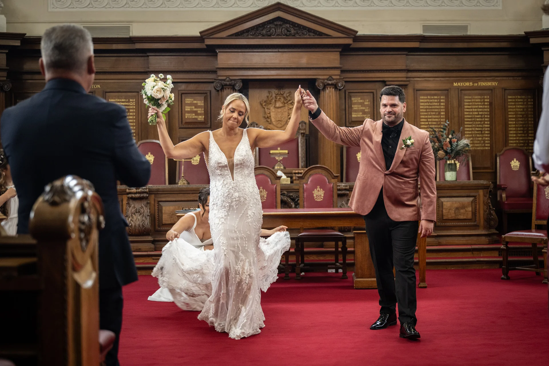 Newlyweds walk hand in hand down the red carpet of a historic town hall chamber with wood panelling, as a guest adjusts the bride's lace wedding dress.