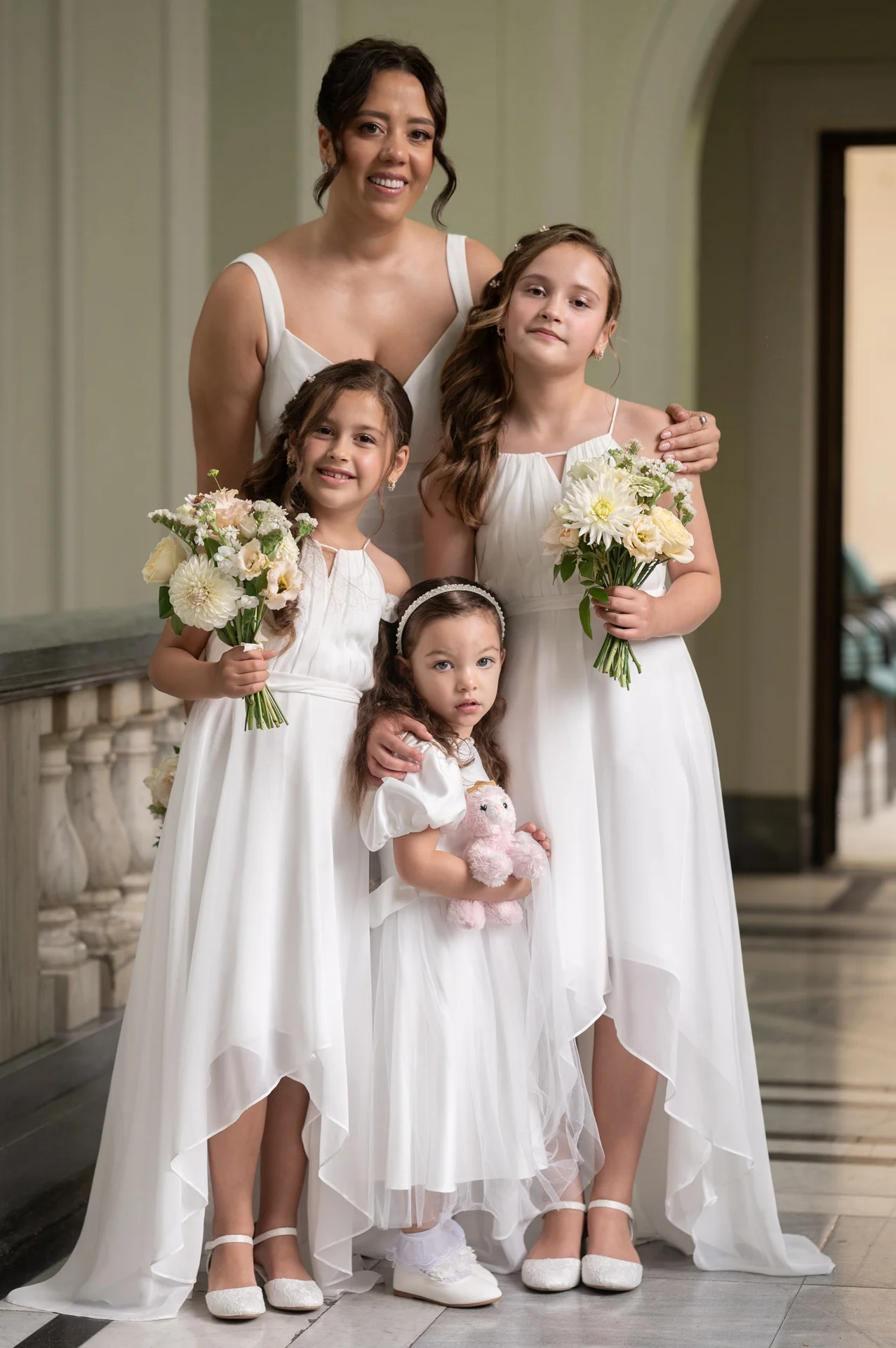 An adult woman and three young flower girls in matching white dresses pose together holding small bouquets inside a classic venue.