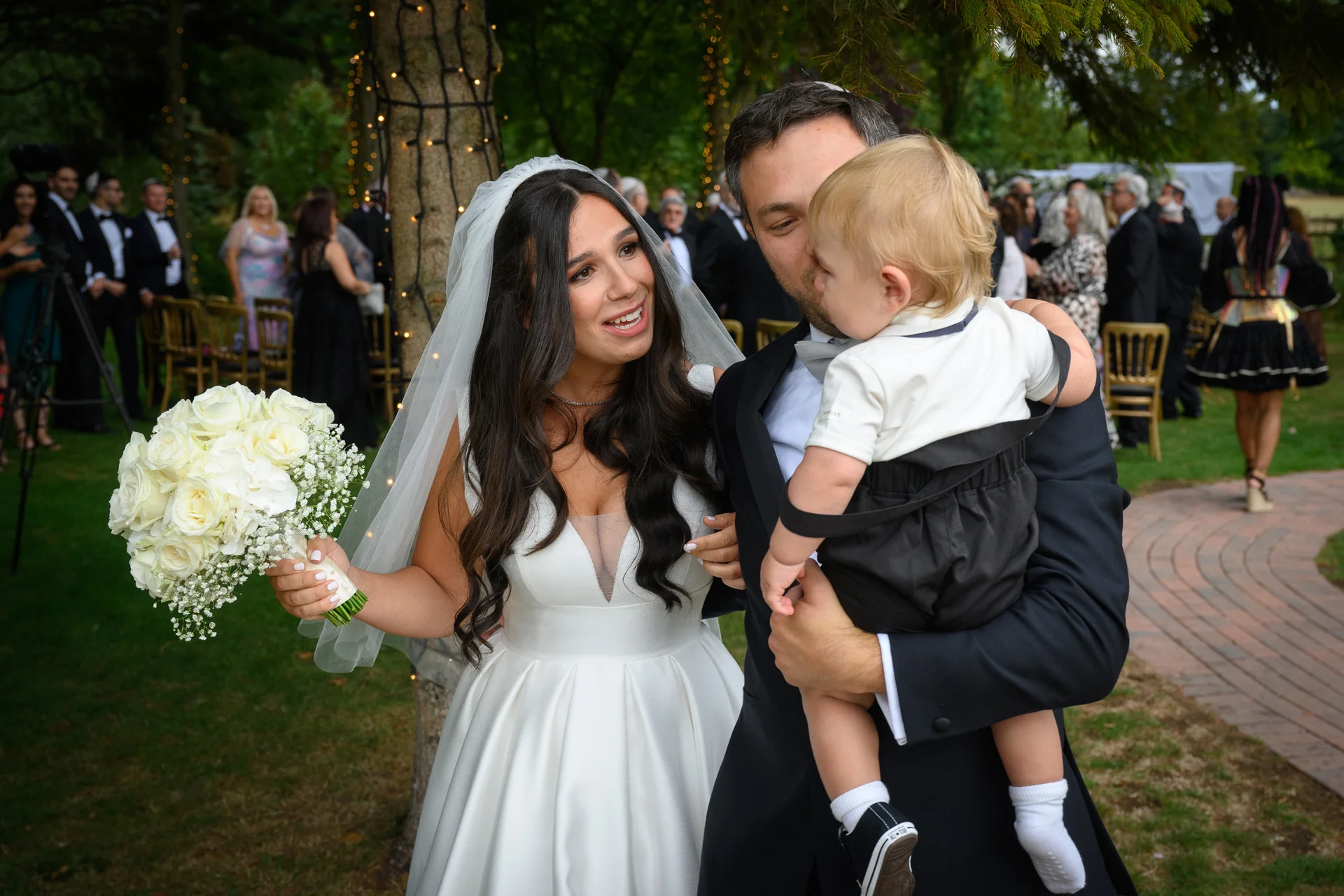 A smiling bride holding a white rose bouquet stands alongside a groom who is holding a young child outdoors during a wedding reception.