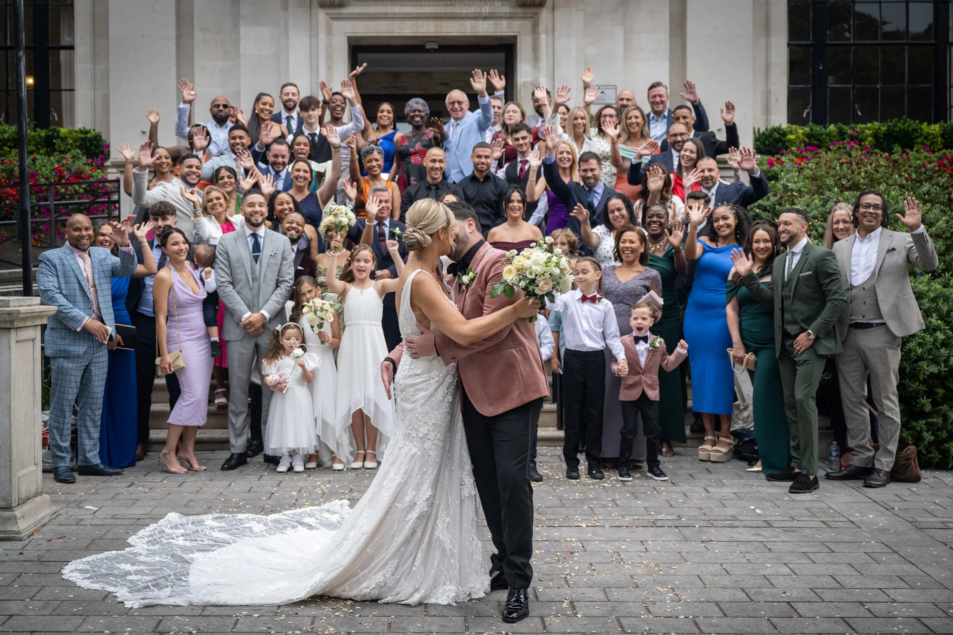 A newlywed couple shares a kiss in the foreground while a large crowd of wedding guests waves and cheers from the stone steps of a grand venue behind them.
