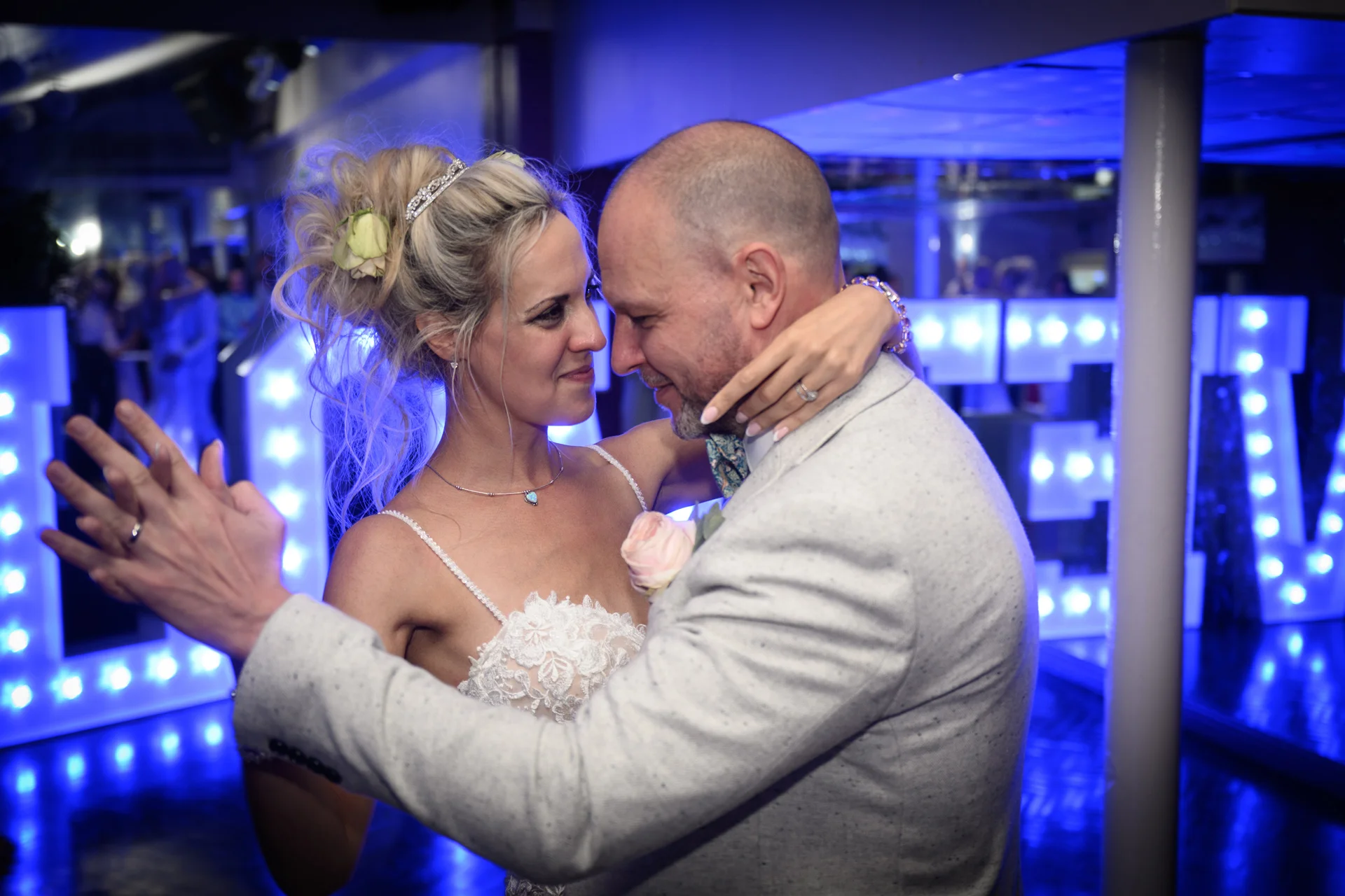 A newlywed couple shares an intimate first dance together under dramatic blue lighting, with illuminated letters glowing in the background.