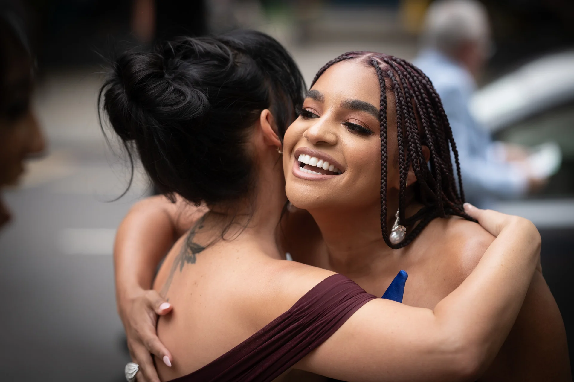Two smiling brides share a warm, joyful embrace outdoors in a city setting, with one bride showcasing intricate box braids and elegant pearl drop earrings.