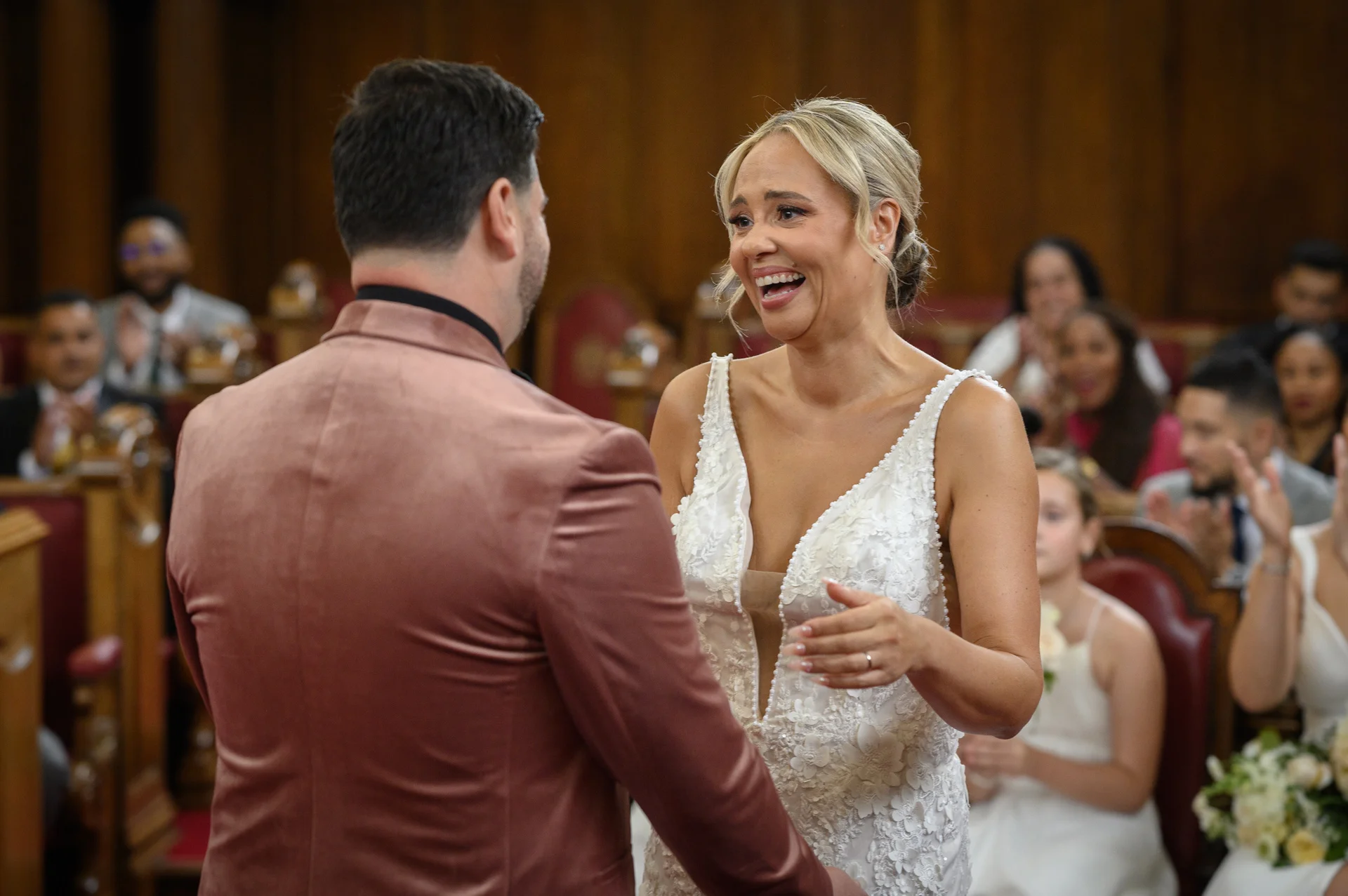 A newlywed bride laughs joyfully as she faces her groom wearing a pink velvet jacket during their ceremony inside a wood-panelled venue.