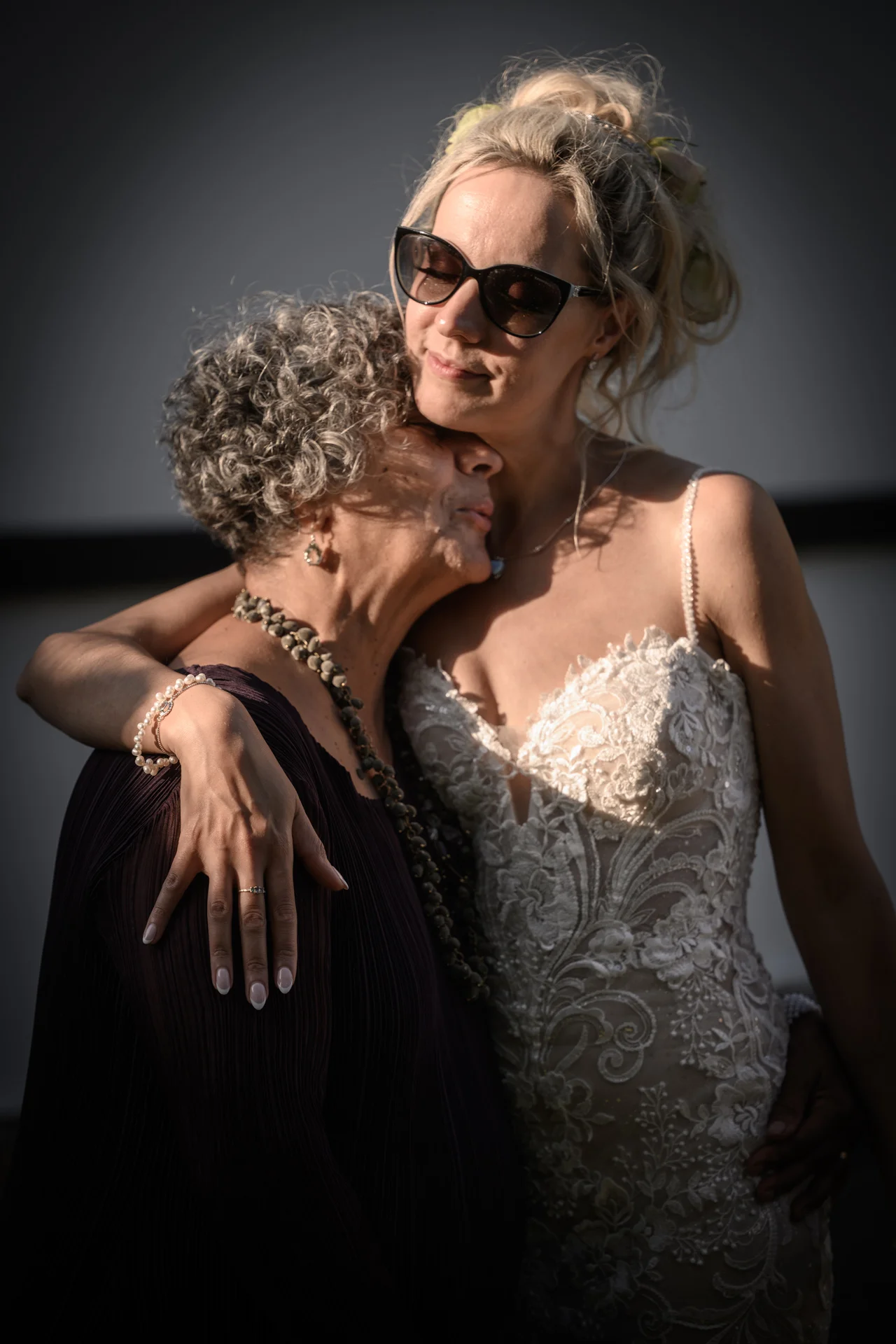 A mature bride wearing a lace wedding dress and sunglasses lovingly embraces an older guest in a tender moment bathed in warm golden hour light.