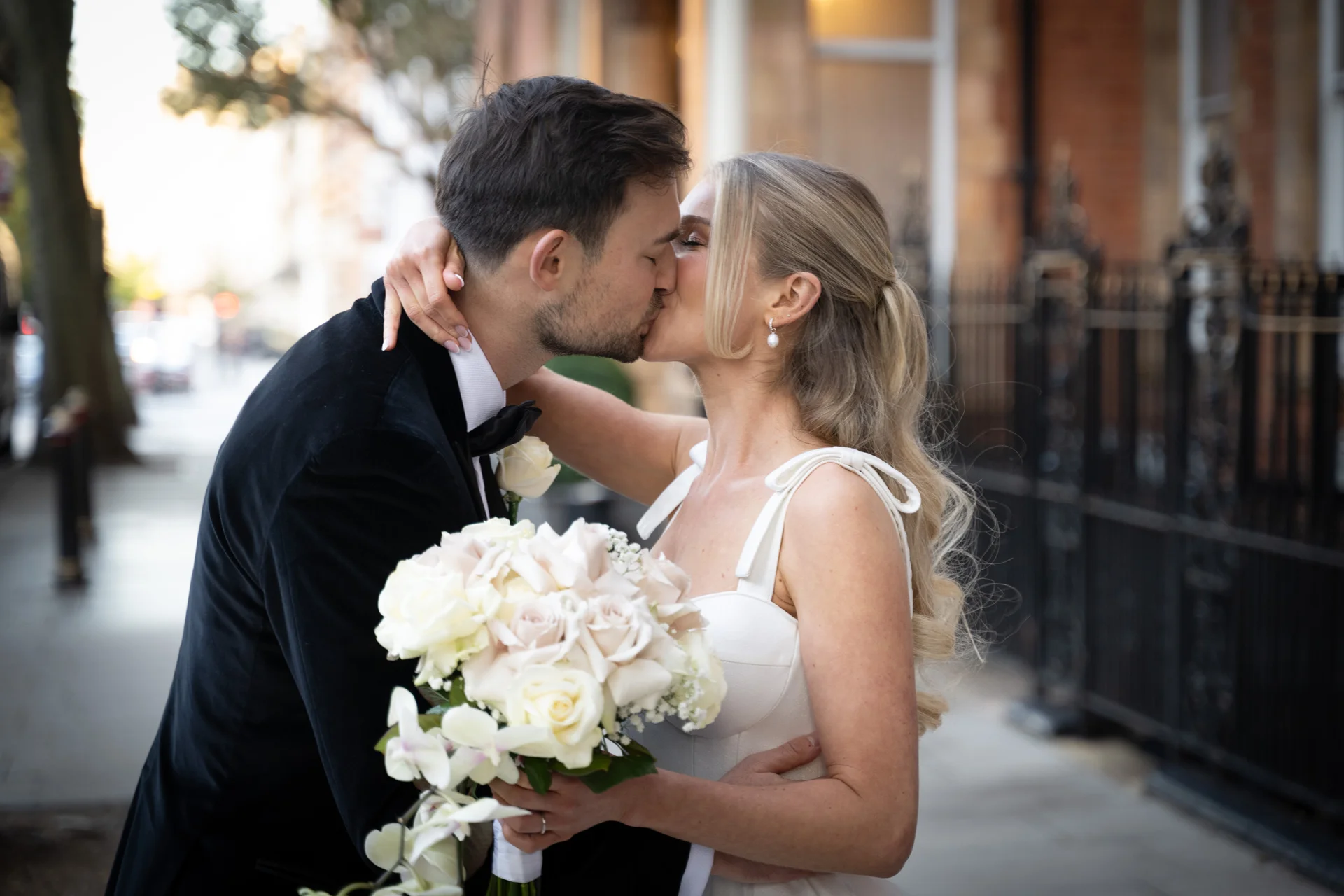 A bride and groom share a tender kiss on a city street, with the bride holding a lush bouquet of white roses and orchids.