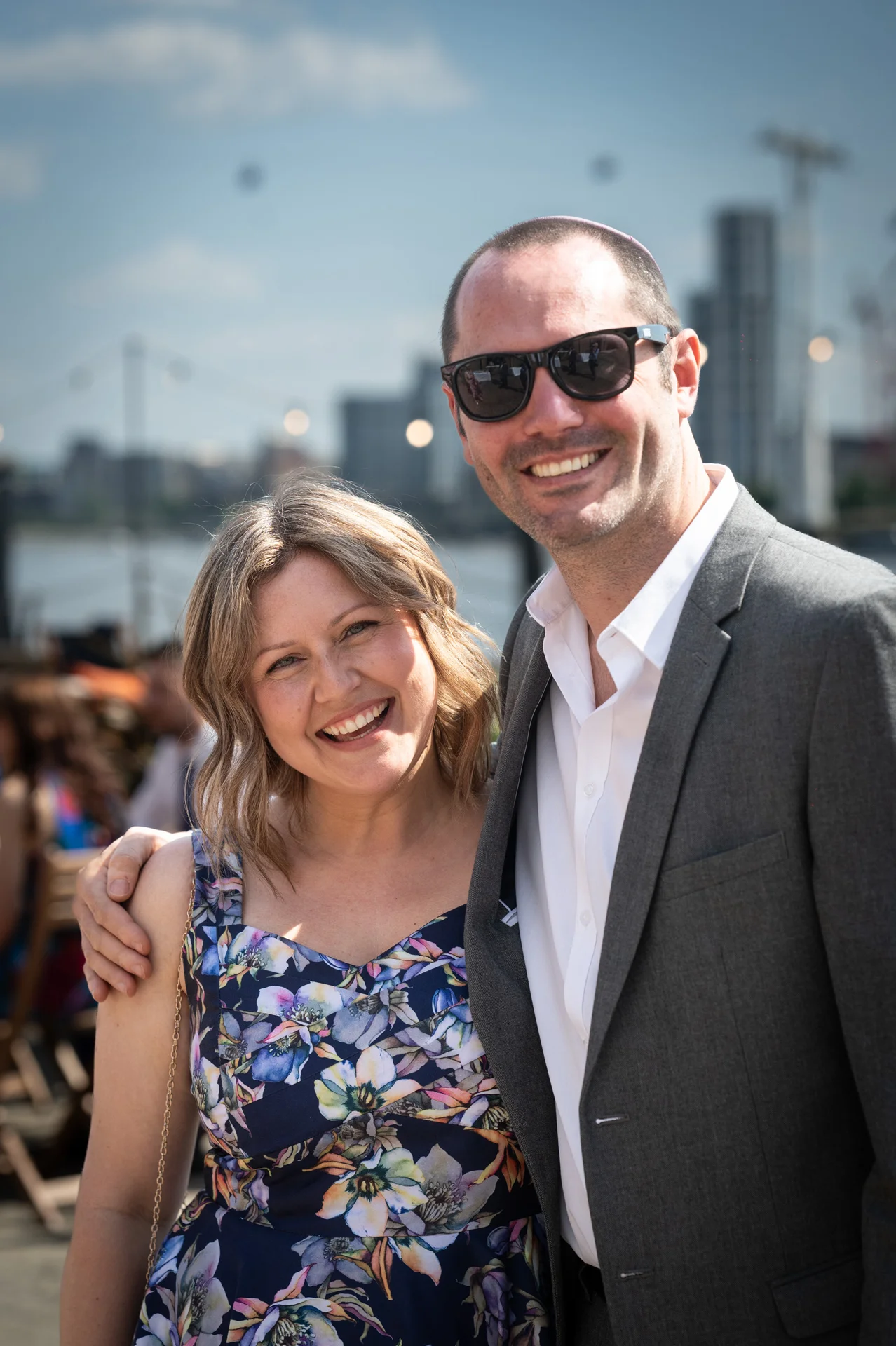 A smiling wedding guest in a floral print dress and a man in a grey suit and sunglasses pose together outdoors in the sunshine.