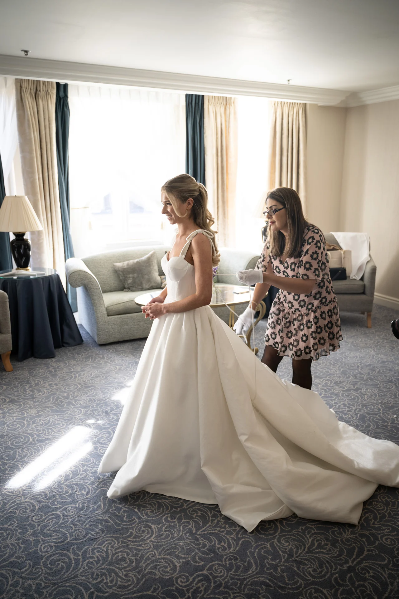 A bride in a structured satin A-line gown with a dramatic train stands in a hotel room as final adjustments are made to her dress.