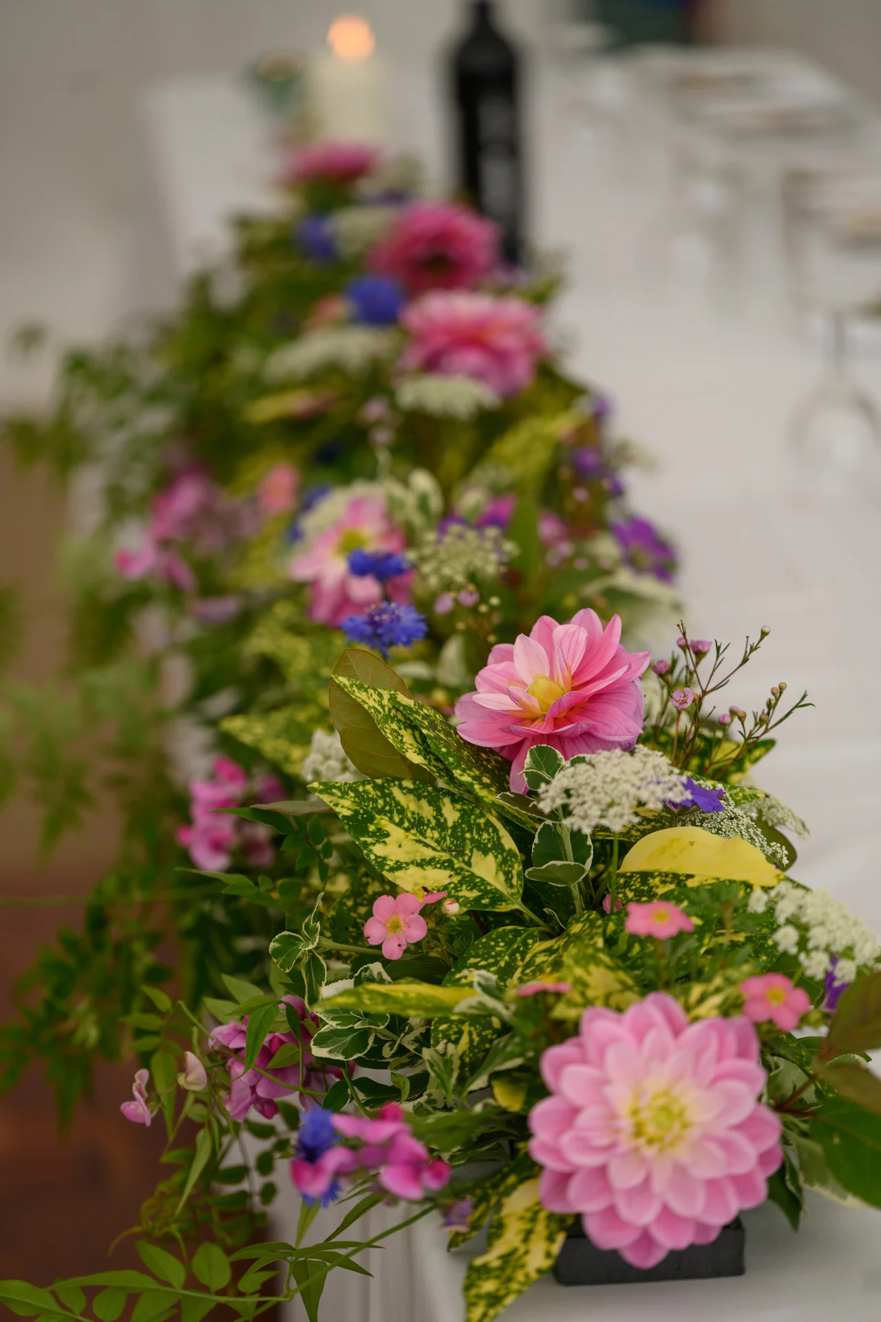 A lush, vibrant pink floral garland runner adorned with dahlias and variegated foliage stretches down the length of a white-clothed wedding banquet table.