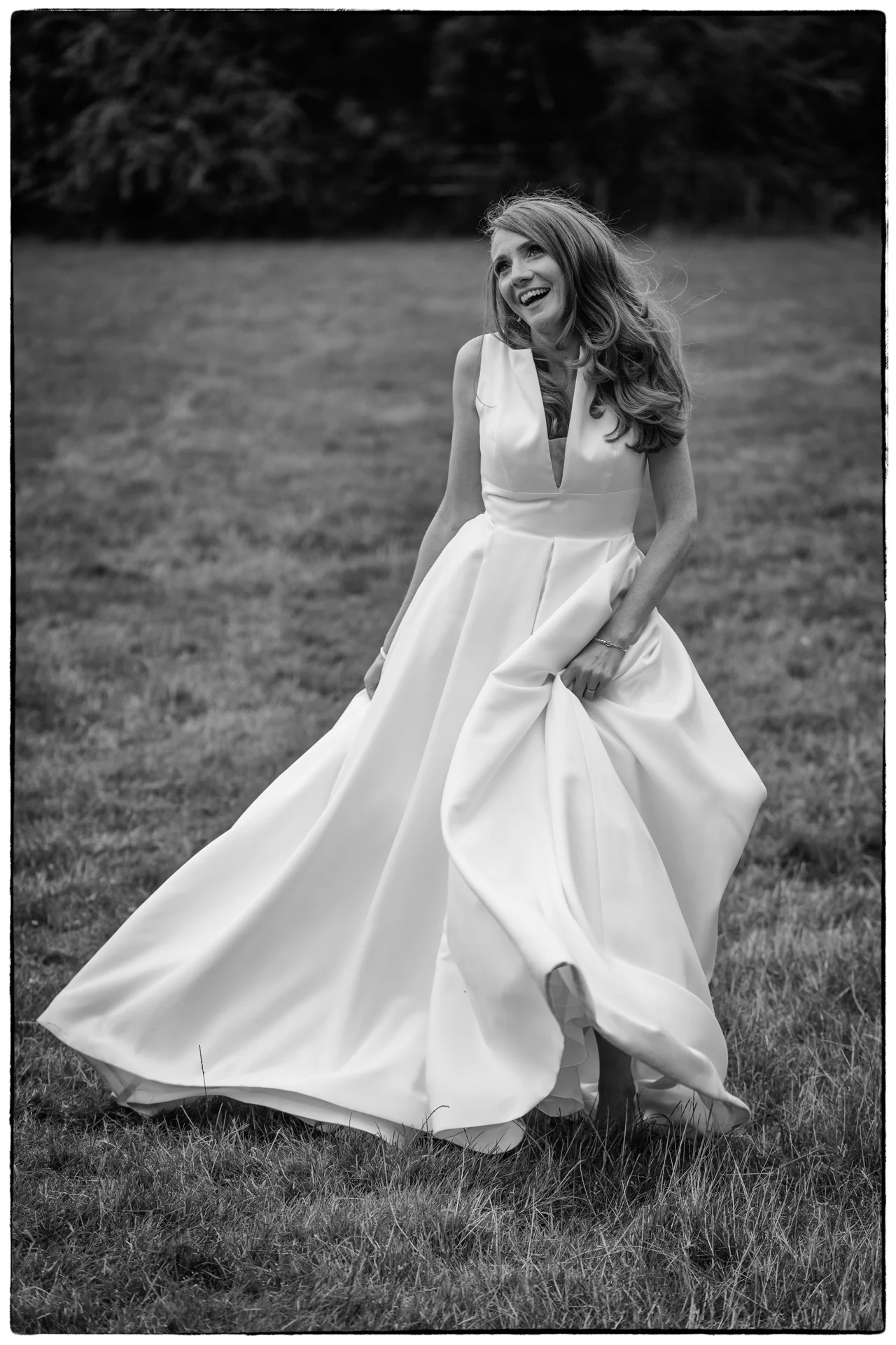 A joyful bride in a white satin A-line gown laughs and twirls through a grassy field in this black and white wedding photograph.