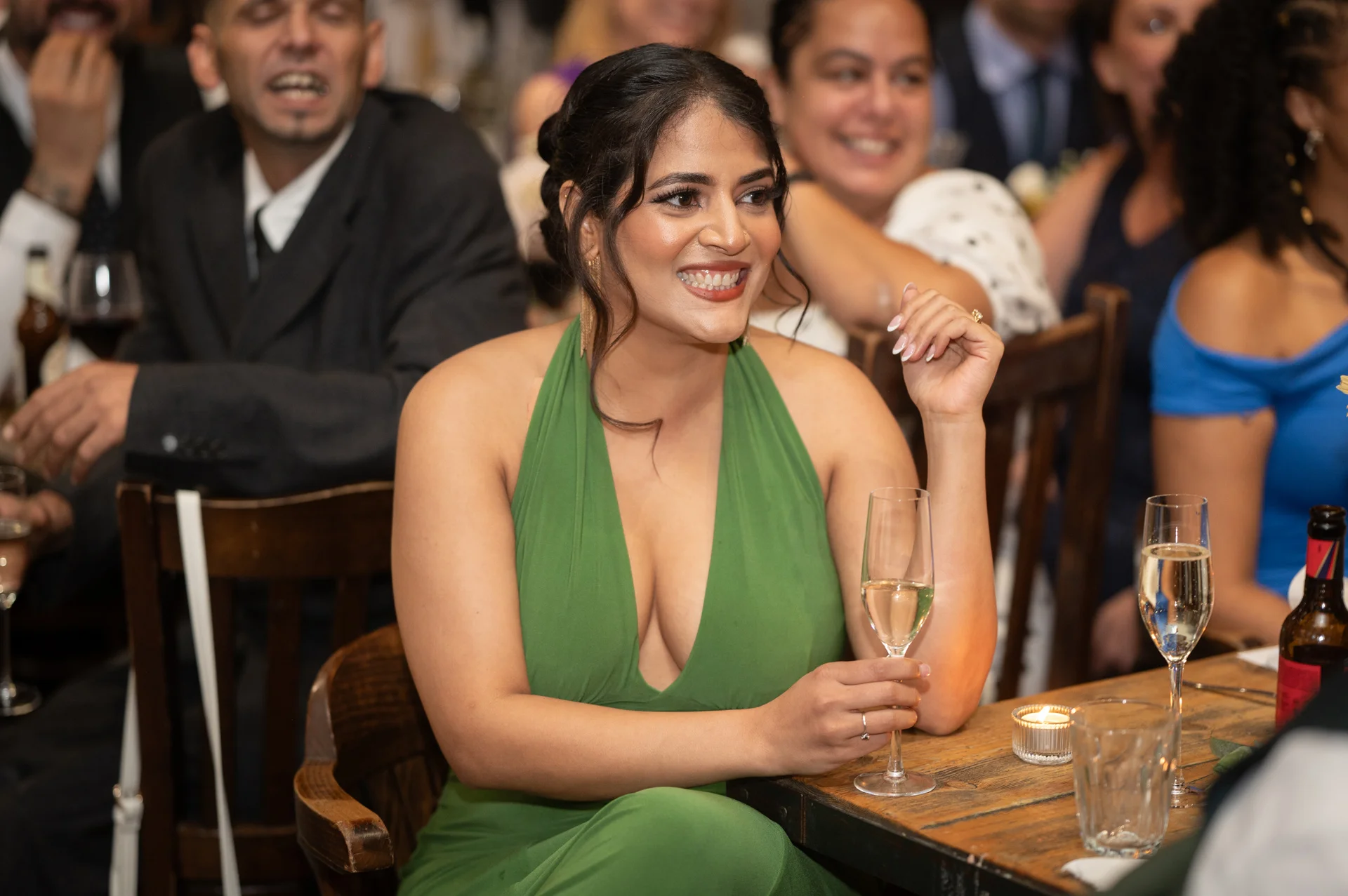A smiling guest wearing a green halter-neck dress sits at a wooden banquet table during the wedding reception, holding a glass of champagne while watching the speeches.
