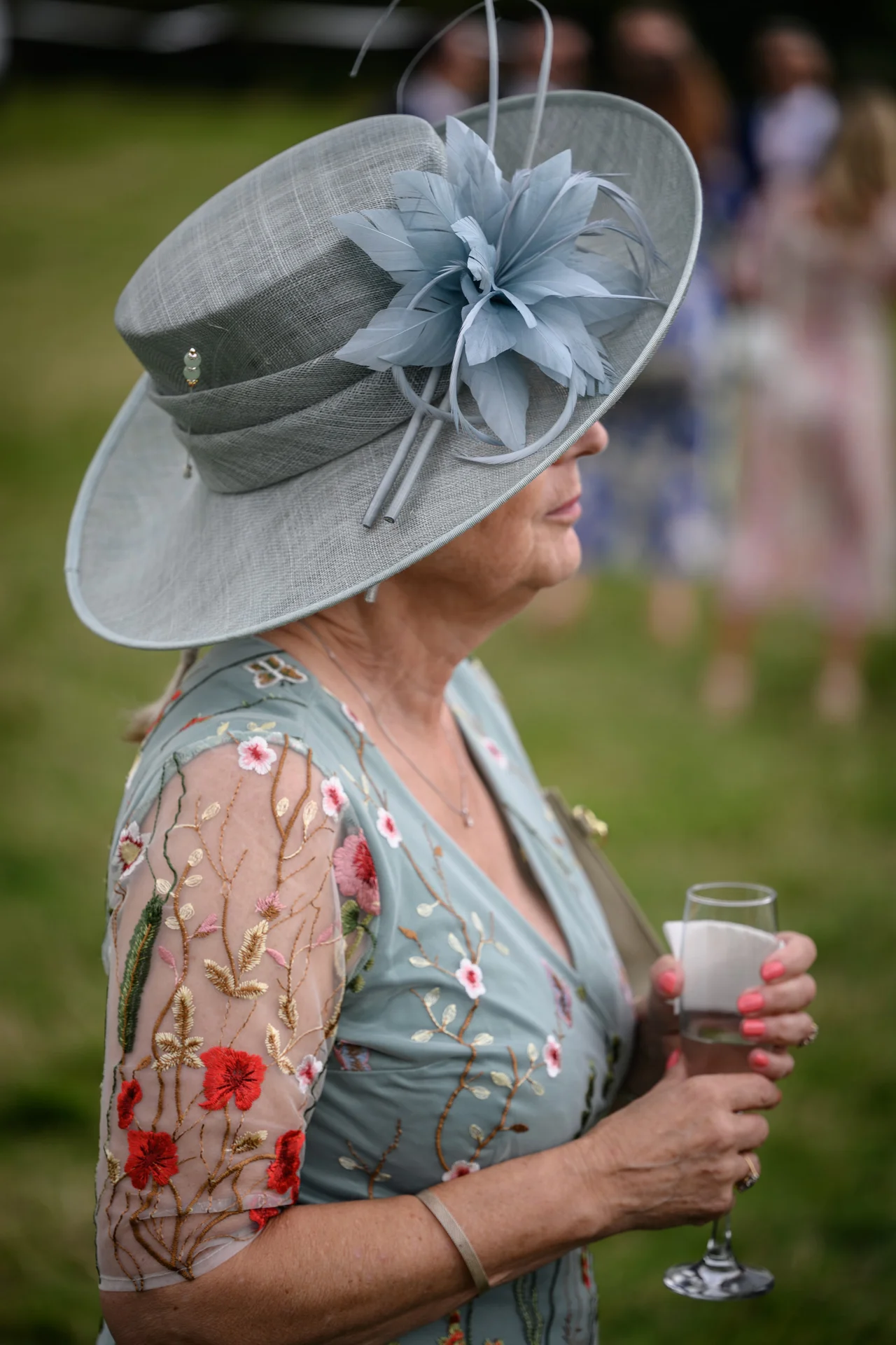 A wedding guest wearing a wide-brimmed sage hat with feather detailing and a floral embroidered dress holds a glass of champagne outdoors.