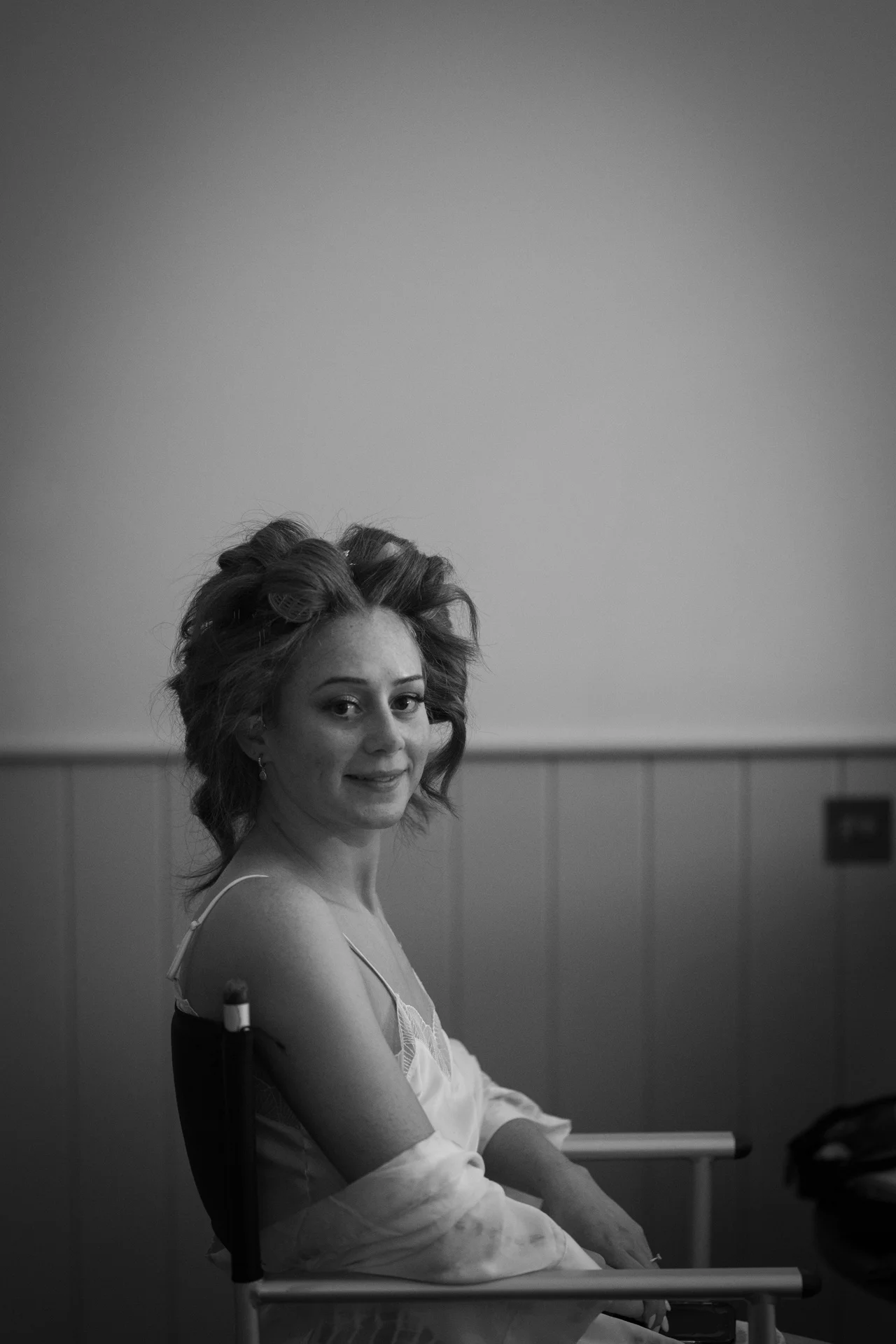 A black and white, over-the-shoulder shot captures a bride sitting in a makeup chair with her hair styled in rollers, glancing back towards the camera with a gentle smile.