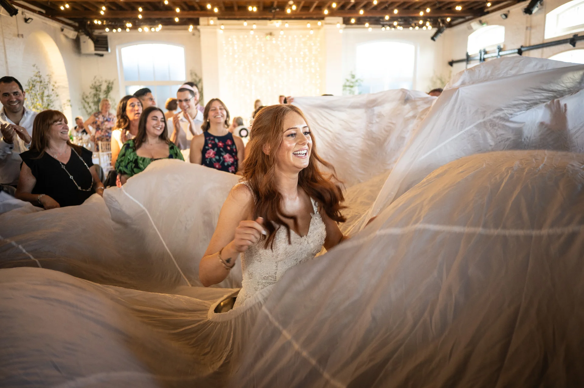 A joyful red-haired bride laughs as she is enveloped underneath an enormous white wedding veil, surrounded by cheering guests in a brightly lit rustic venue.