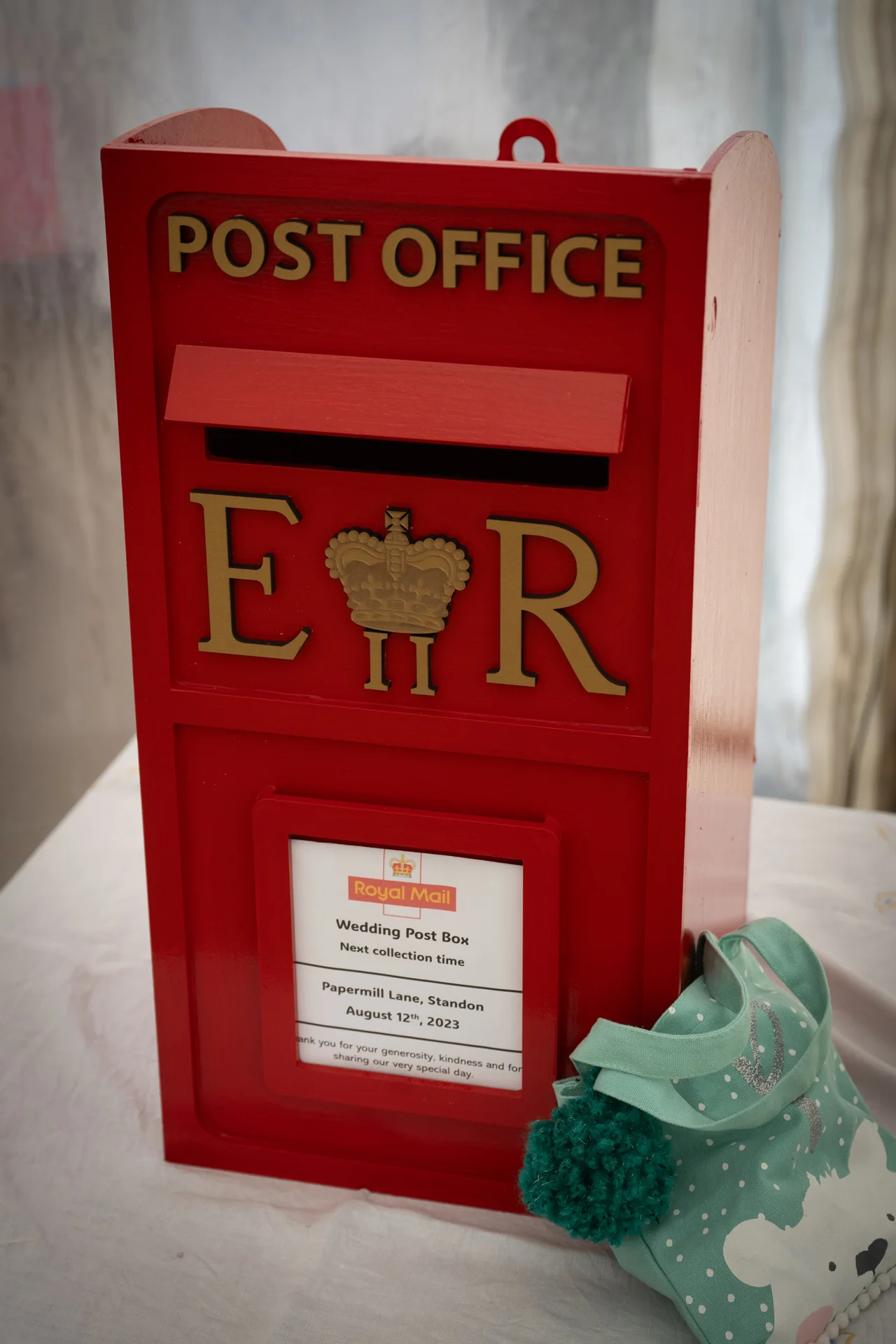 A traditional red post box used as a wedding card box sits on a table alongside a small turquoise patterned bag.
