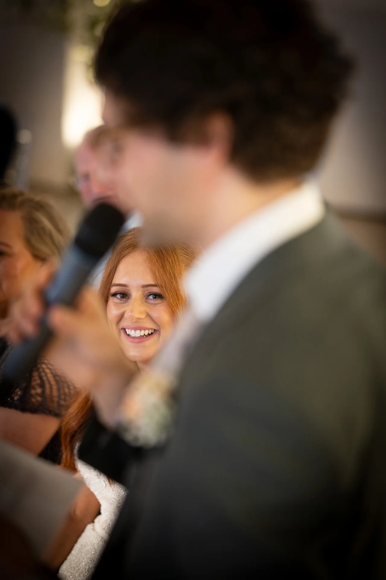 A smiling bride with red hair looks on happily as someone delivers a speech during the wedding reception, captured through a blurred foreground.