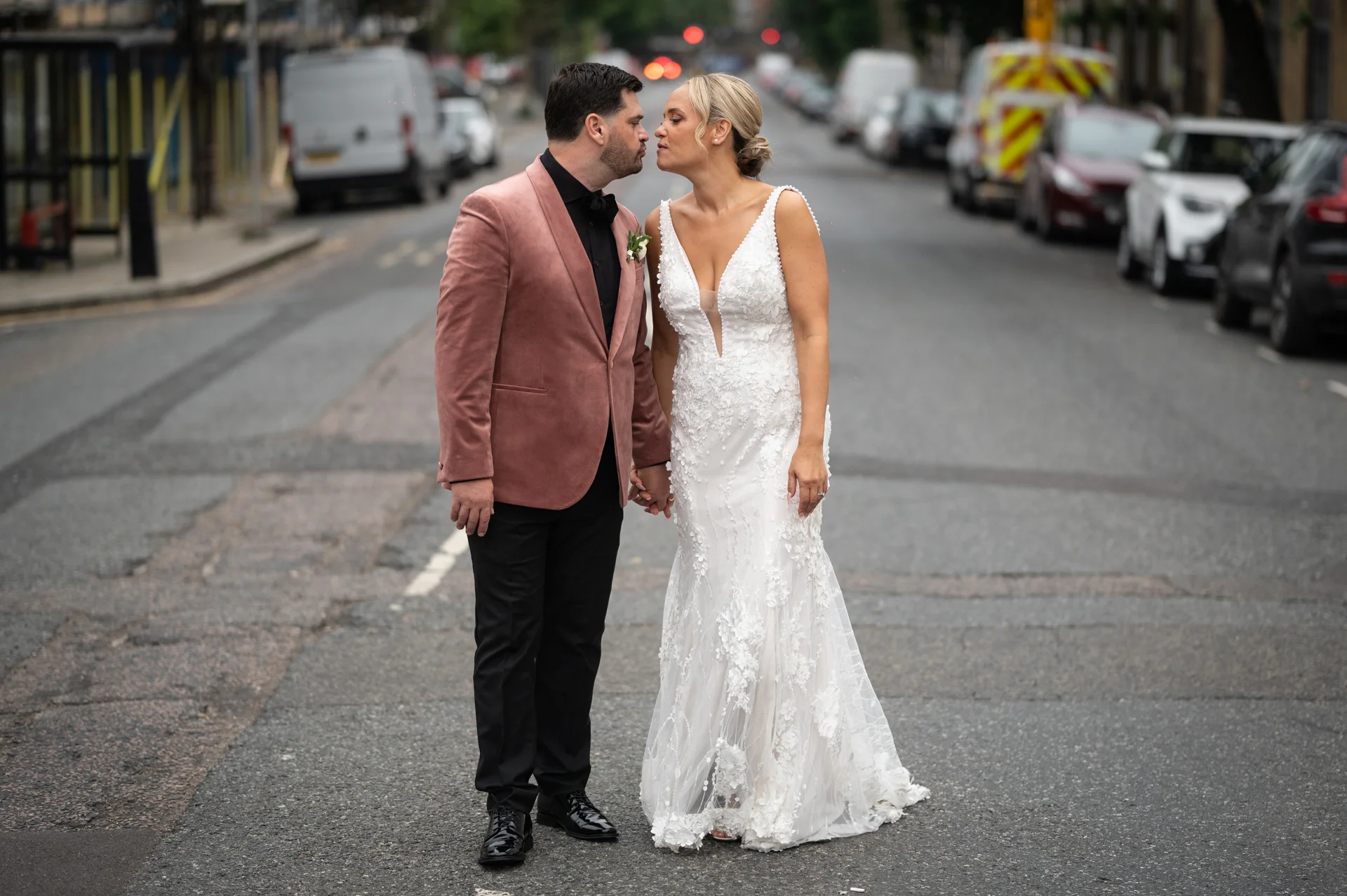 A newlywed couple shares a tender kiss while holding hands in the middle of a bustling city street during their urban wedding celebration.