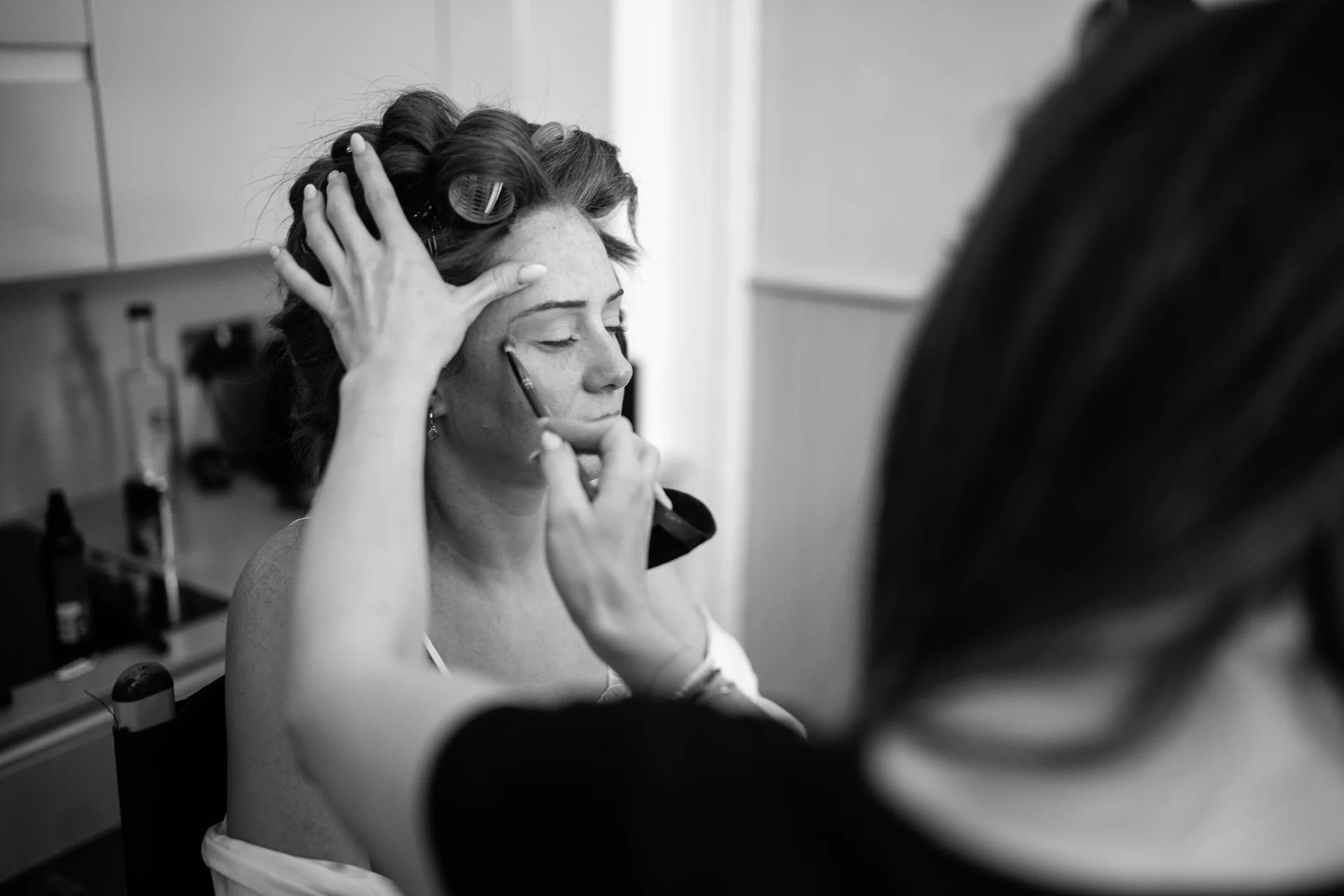 A candid black and white over-the-shoulder shot captures a bride having her makeup applied with rollers in her hair during morning bridal preparations.