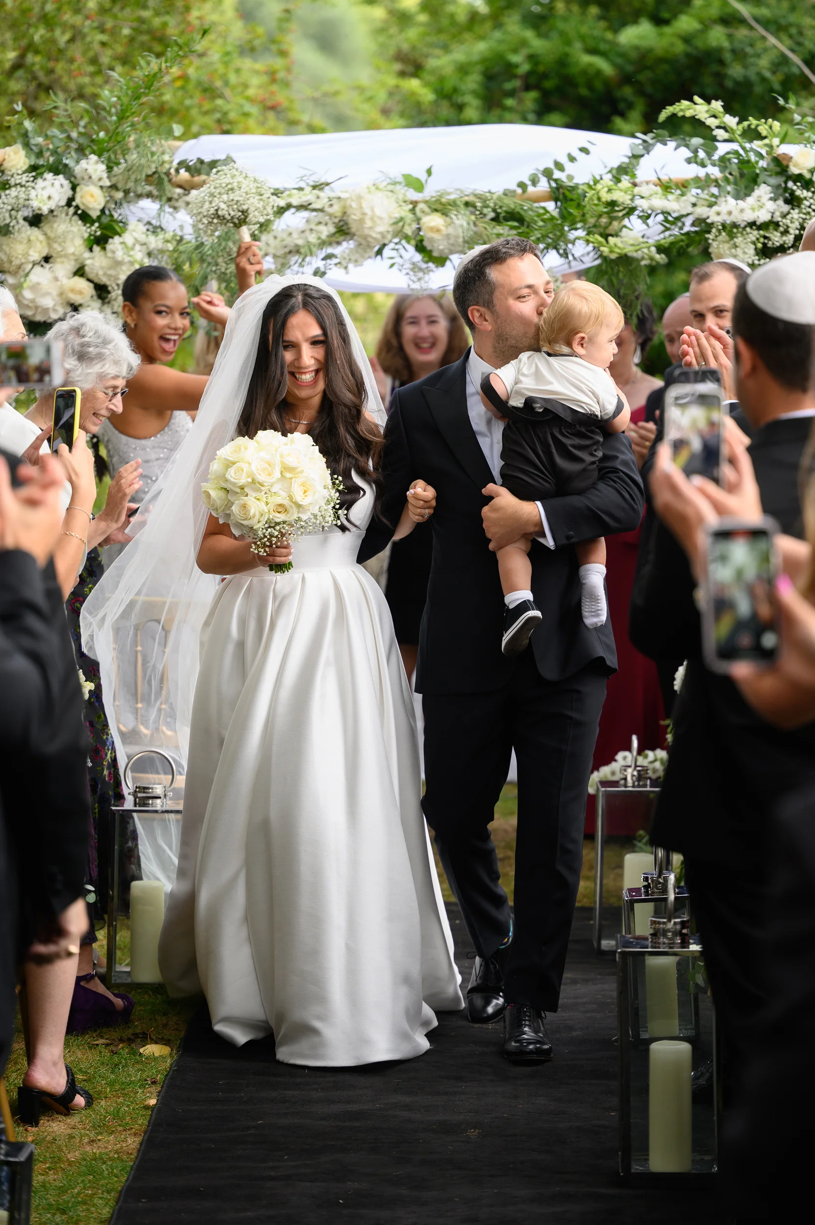 A joyous bride and groom walk hand in hand down the aisle under a white chuppah while carrying a young child and smiling guests capture the moment on their phones.