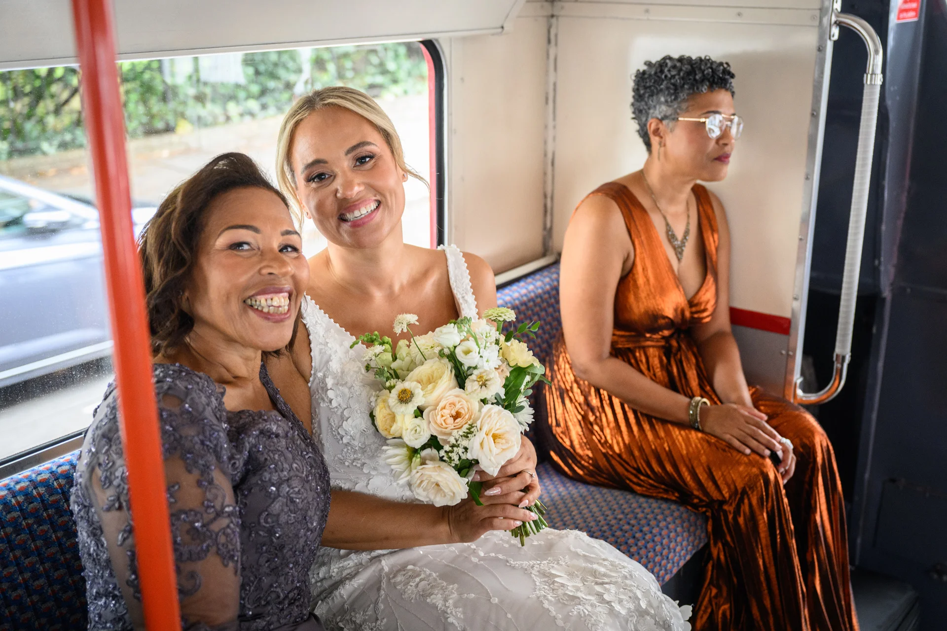 A smiling bride holding a floral bouquet sits on a London bus alongside a family member and a guest.