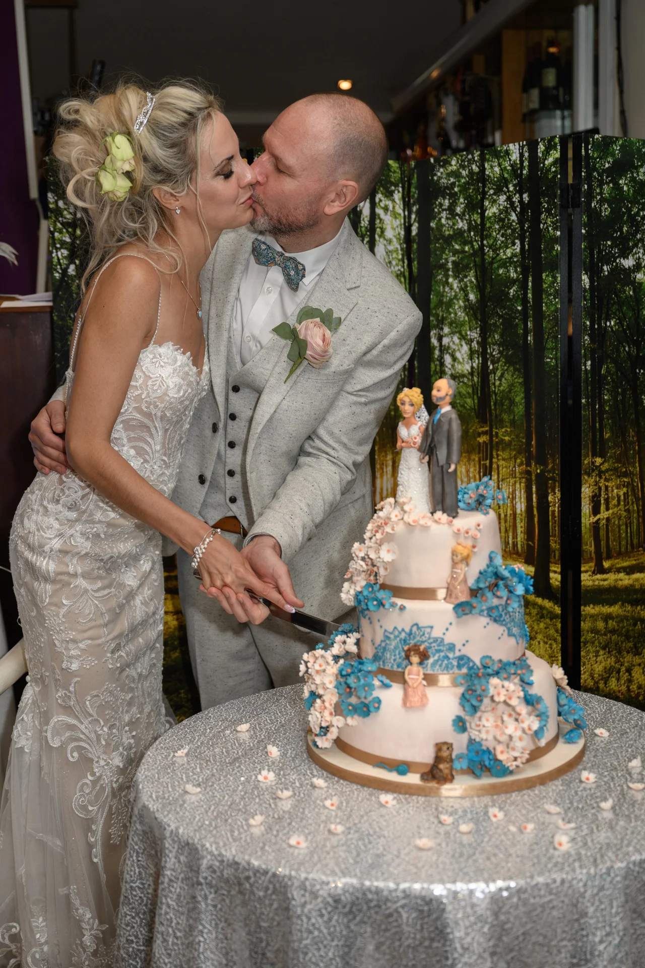 The bride and groom share a tender kiss while cutting their three-tiered wedding cake decorated with blue and pink floral details in front of a woodland backdrop.
