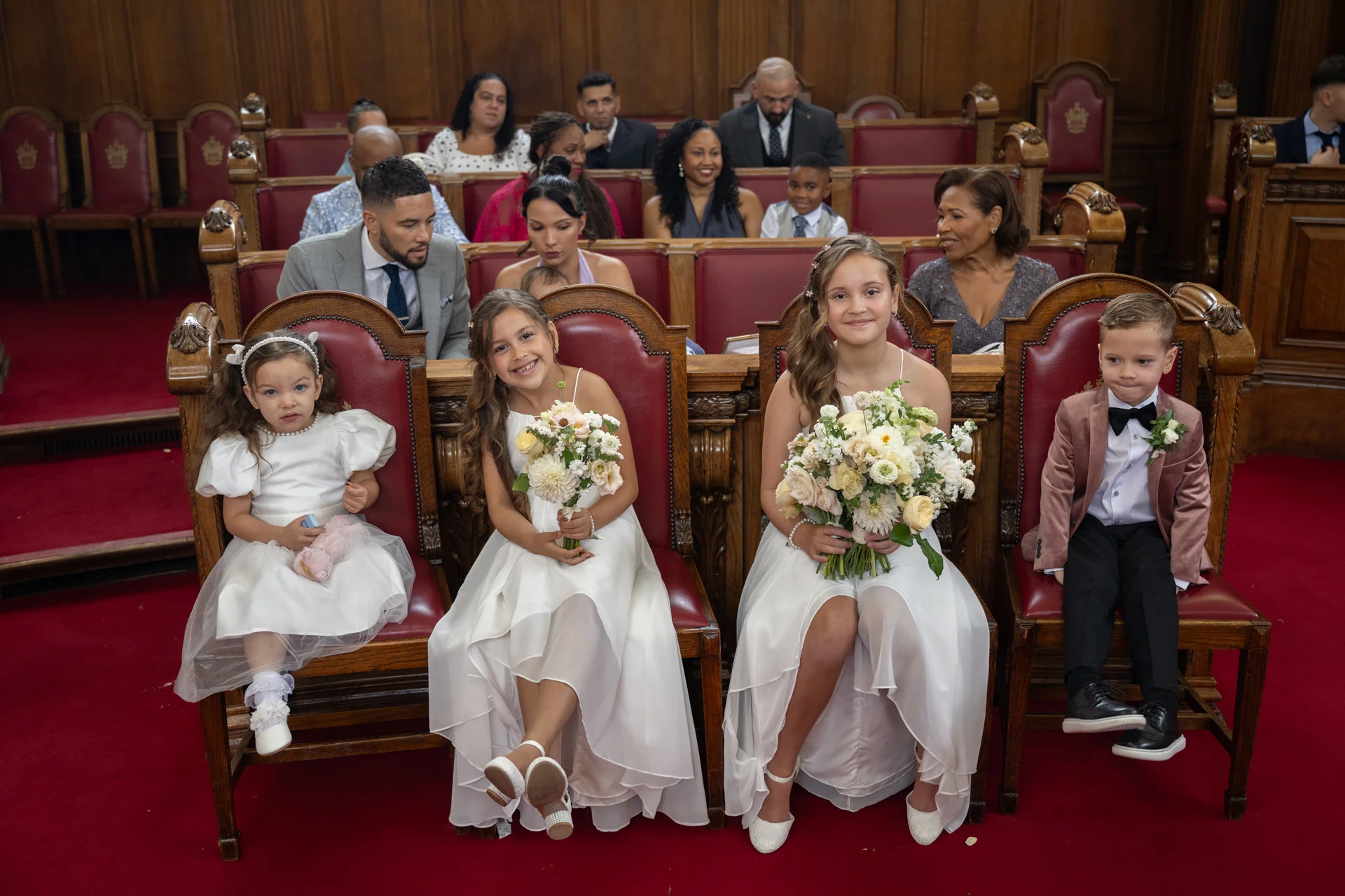Young flower girls holding bouquets and a page boy in a velvet blazer sit smiling on wooden pews during a wedding ceremony.