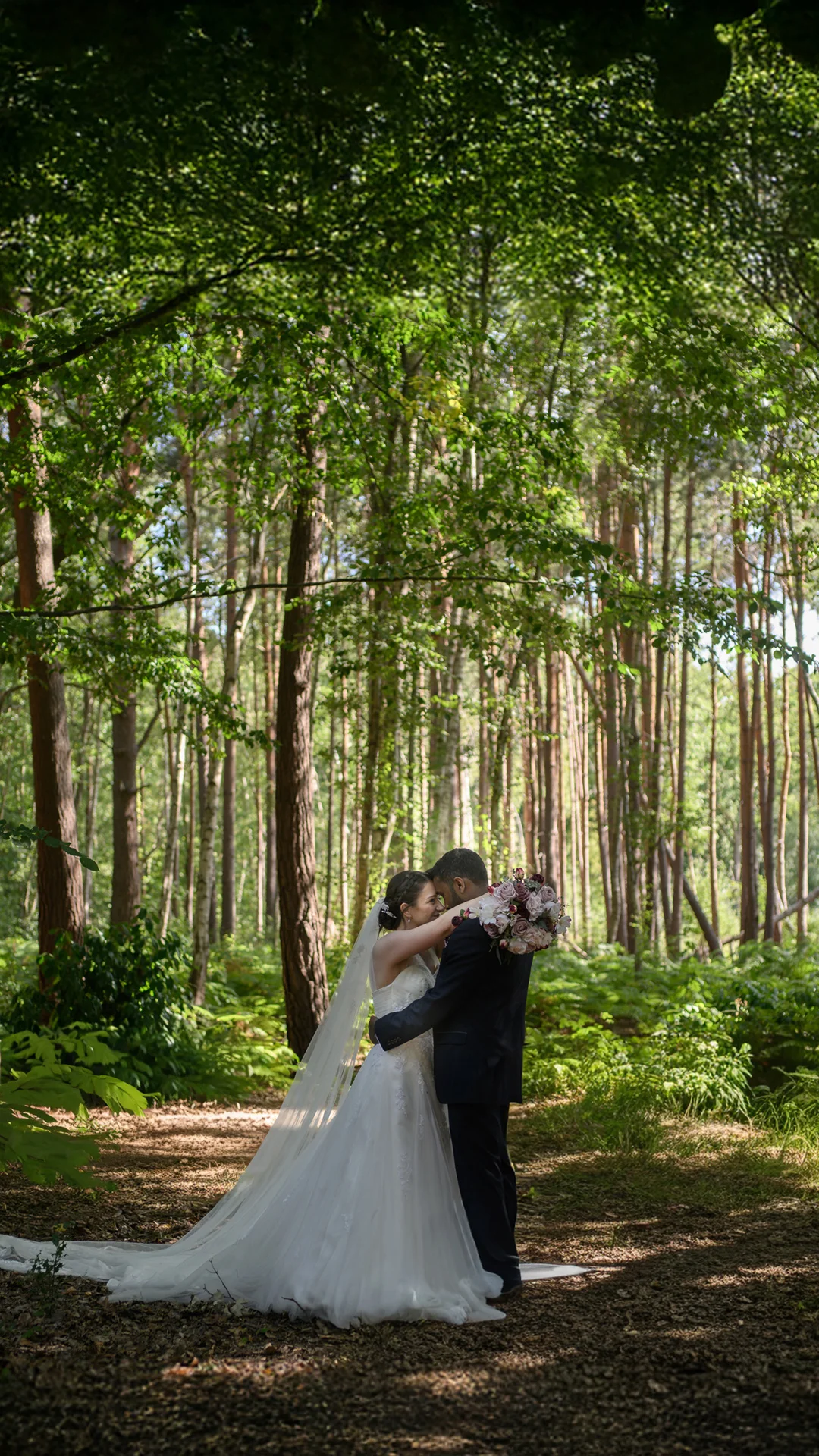 A newlywed couple shares a tender forehead kiss surrounded by a lush green woodland and dappled sunlight.