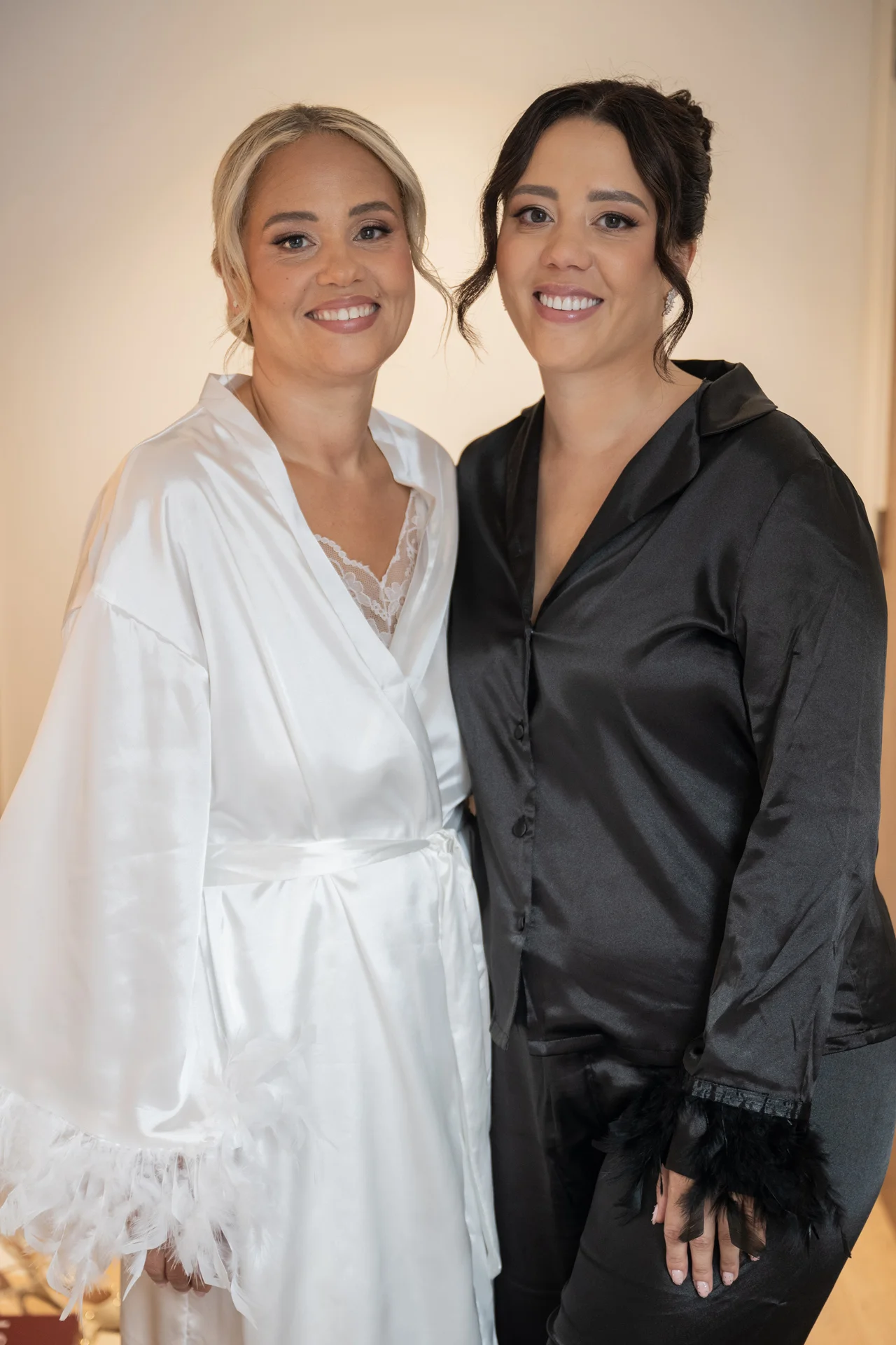 Two smiling brides stand side by side during morning preparations, wearing matching white and black silk robes with feather trim details.