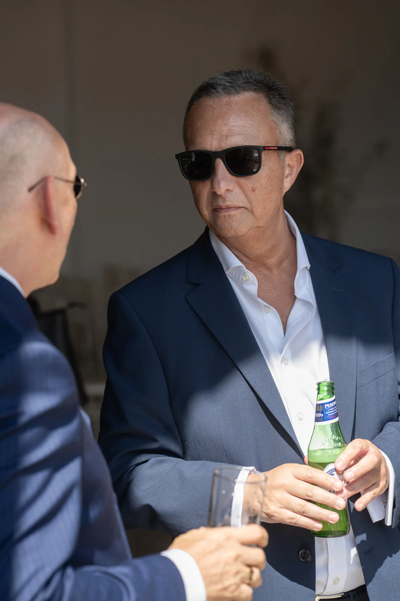 A guest in a navy suit and sunglasses chats and holds a bottle of beer during the wedding drinks reception.