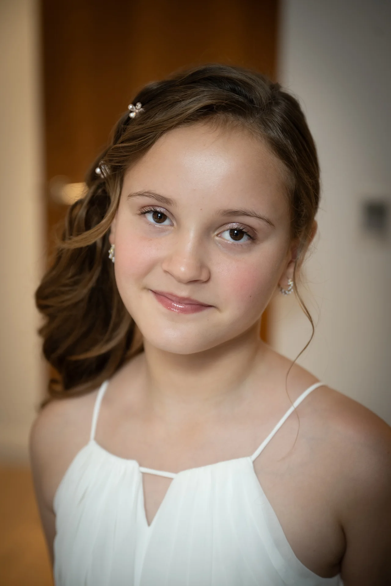 Close-up portrait of a young flower girl wearing a white dress with delicate straps and styled hair adorned with hair pins.