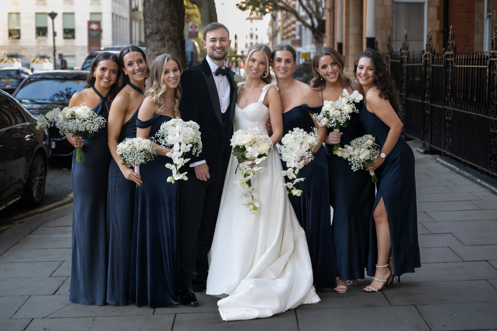The bride in an a-line gown and groom in a black tuxedo pose on a London pavement alongside bridesmaids in matching dark blue velvet and satin dresses holding cascading orchid bouquets.