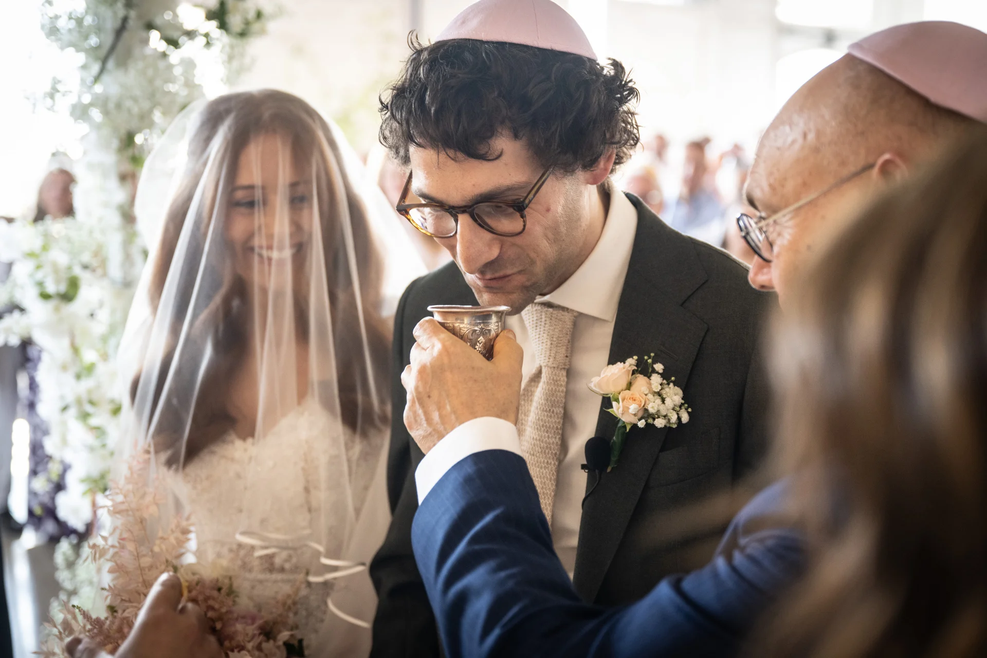 A groom wearing a kippah sips from a silver goblet during a Jewish wedding ceremony while the smiling bride looks on beneath her veil.