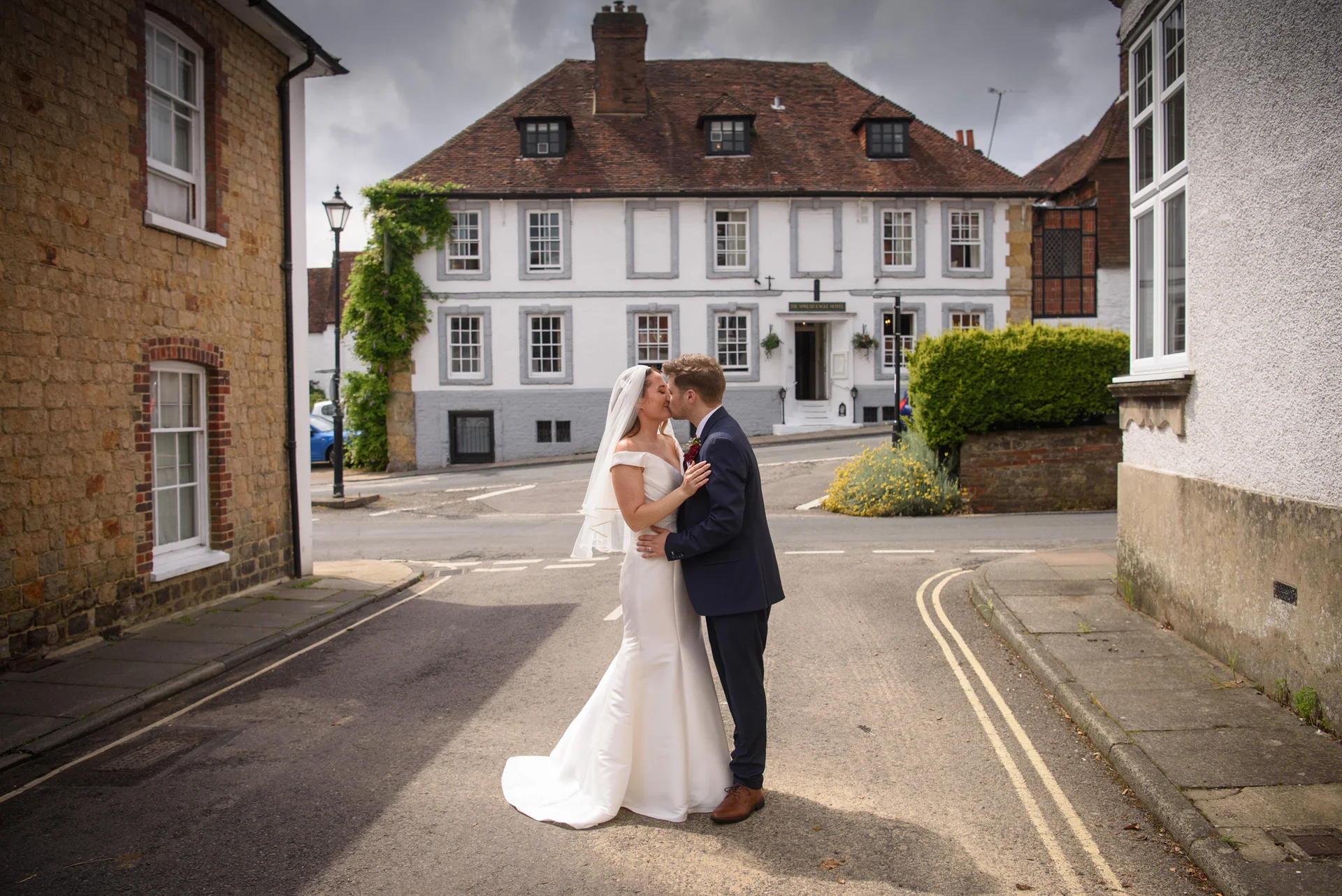 A newlywed couple shares a tender kiss in the middle of a historic town street framed by traditional buildings on an overcast wedding day.