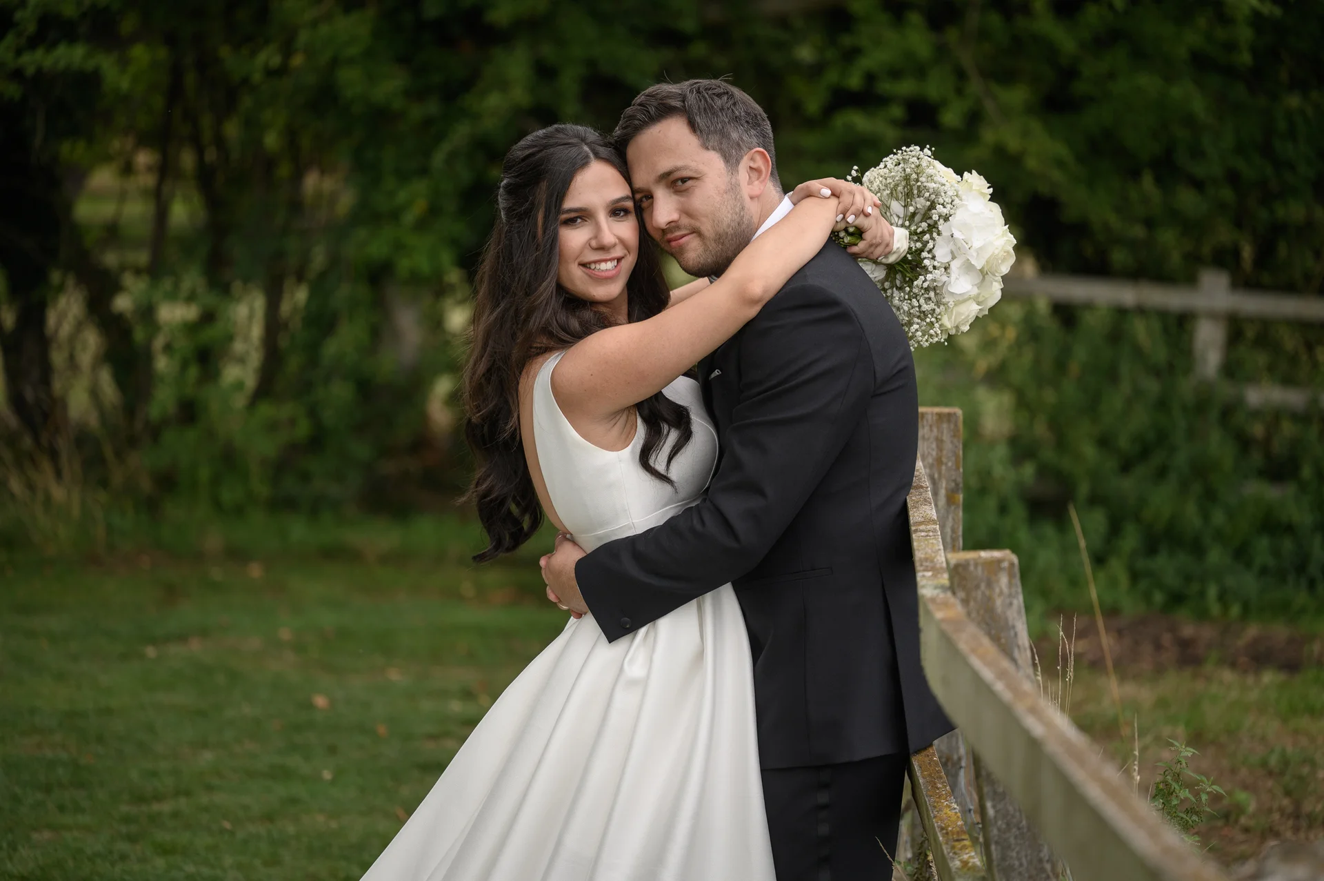 A newly married couple embrace lovingly near a wooden fence outdoors, with the bride holding a white bouquet and wearing a classic satin gown.