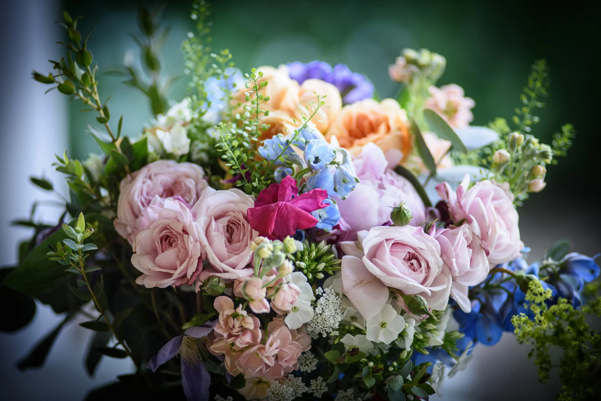 A close-up view of a colourful, hand-tied wedding bouquet featuring pink garden roses, blue delphiniums, and delicate foliage against a soft bokeh background.