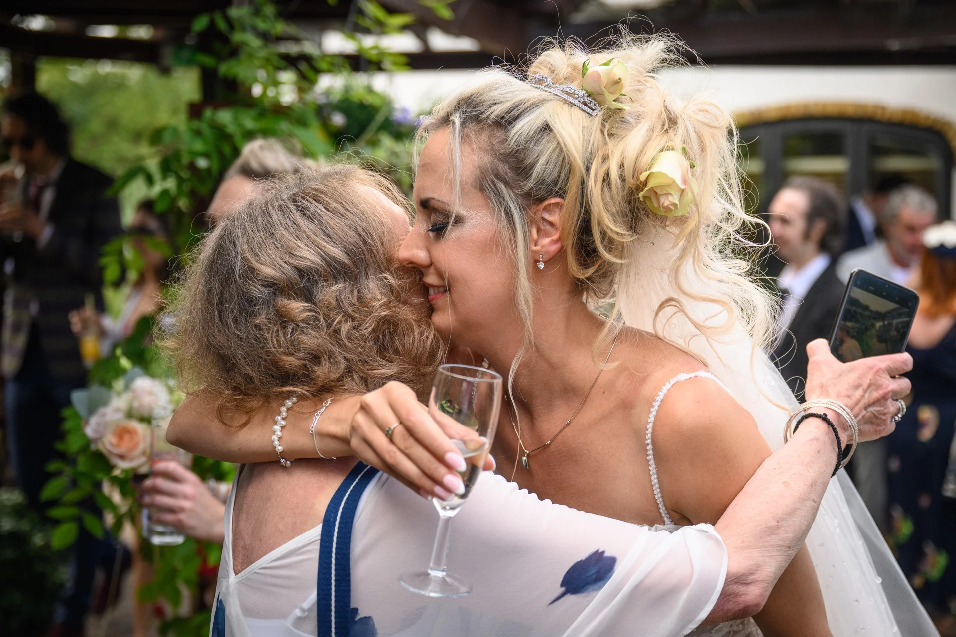 A smiling bride wearing an intricate updo with flowers embraces a wedding guest during a vibrant outdoor drinks reception.