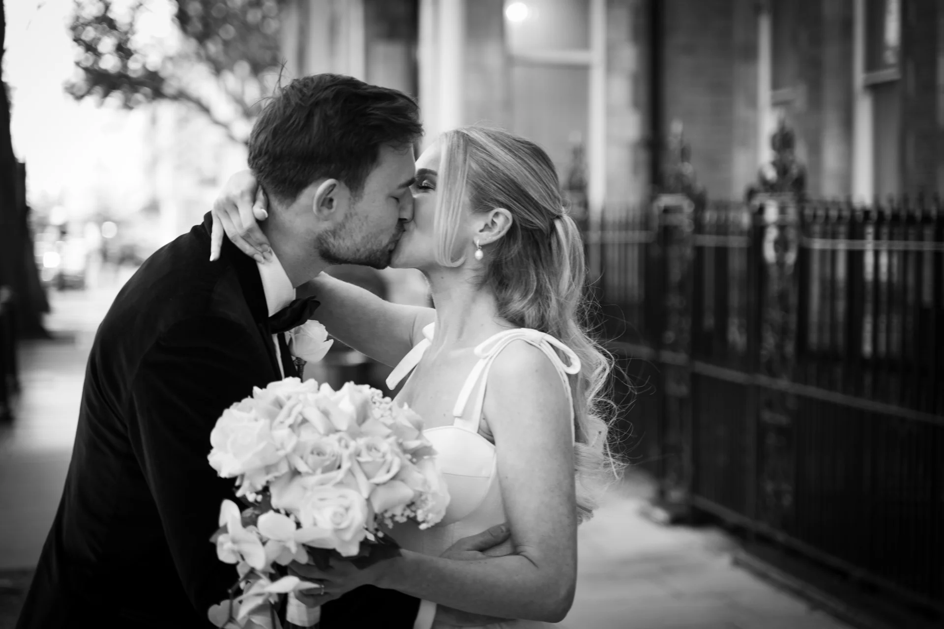 A newlywed couple shares a tender kiss on a London street, the bride holding a white bouquet while wearing a strap dress.