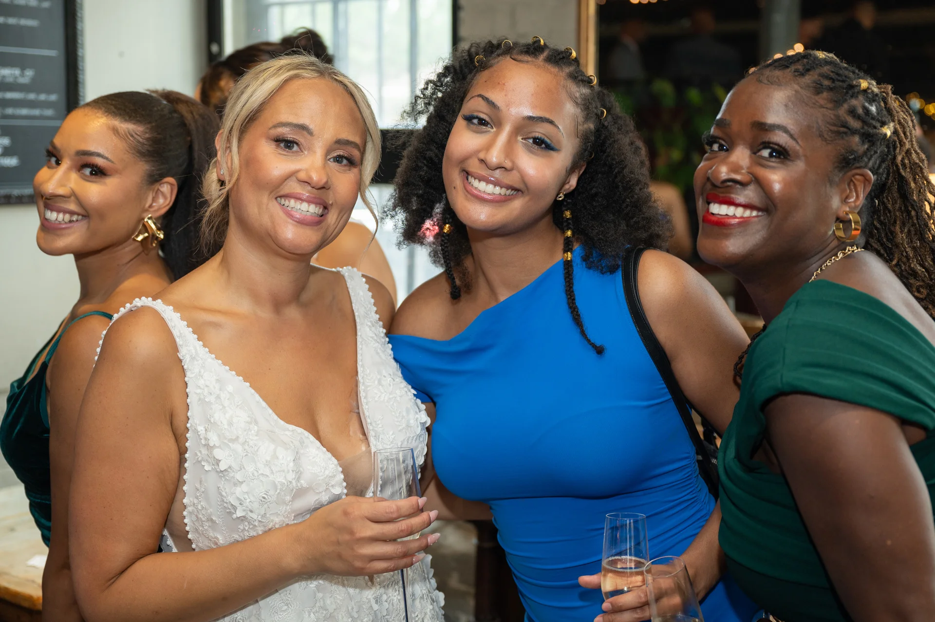 A smiling bride in a lace V-neck wedding dress poses joyfully with her friends holding champagne glasses during the wedding reception.
