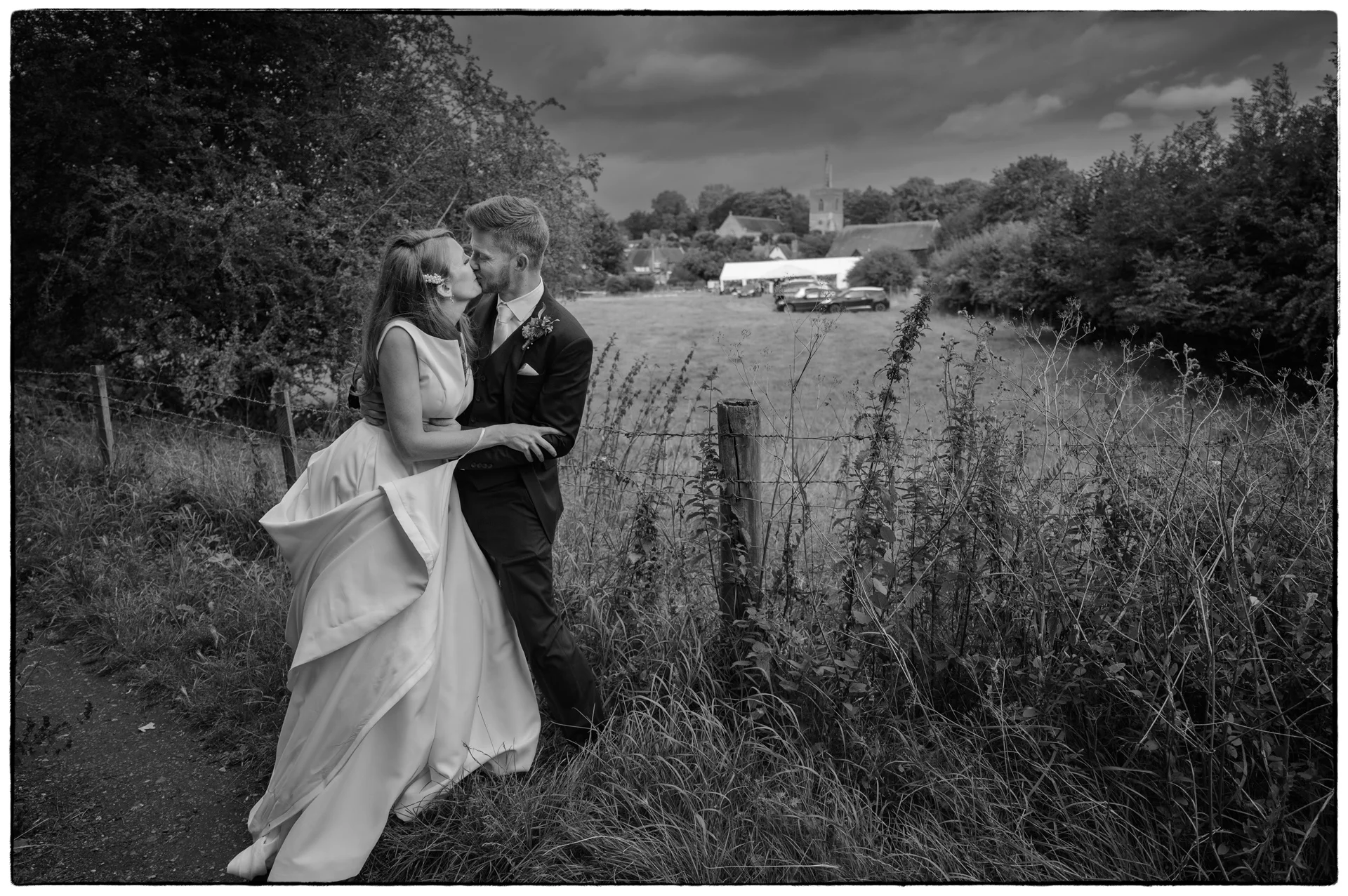 A newlywed couple share a tender kiss along a rural path beside a wooden fence and tall field grass under a dramatic cloudy sky.