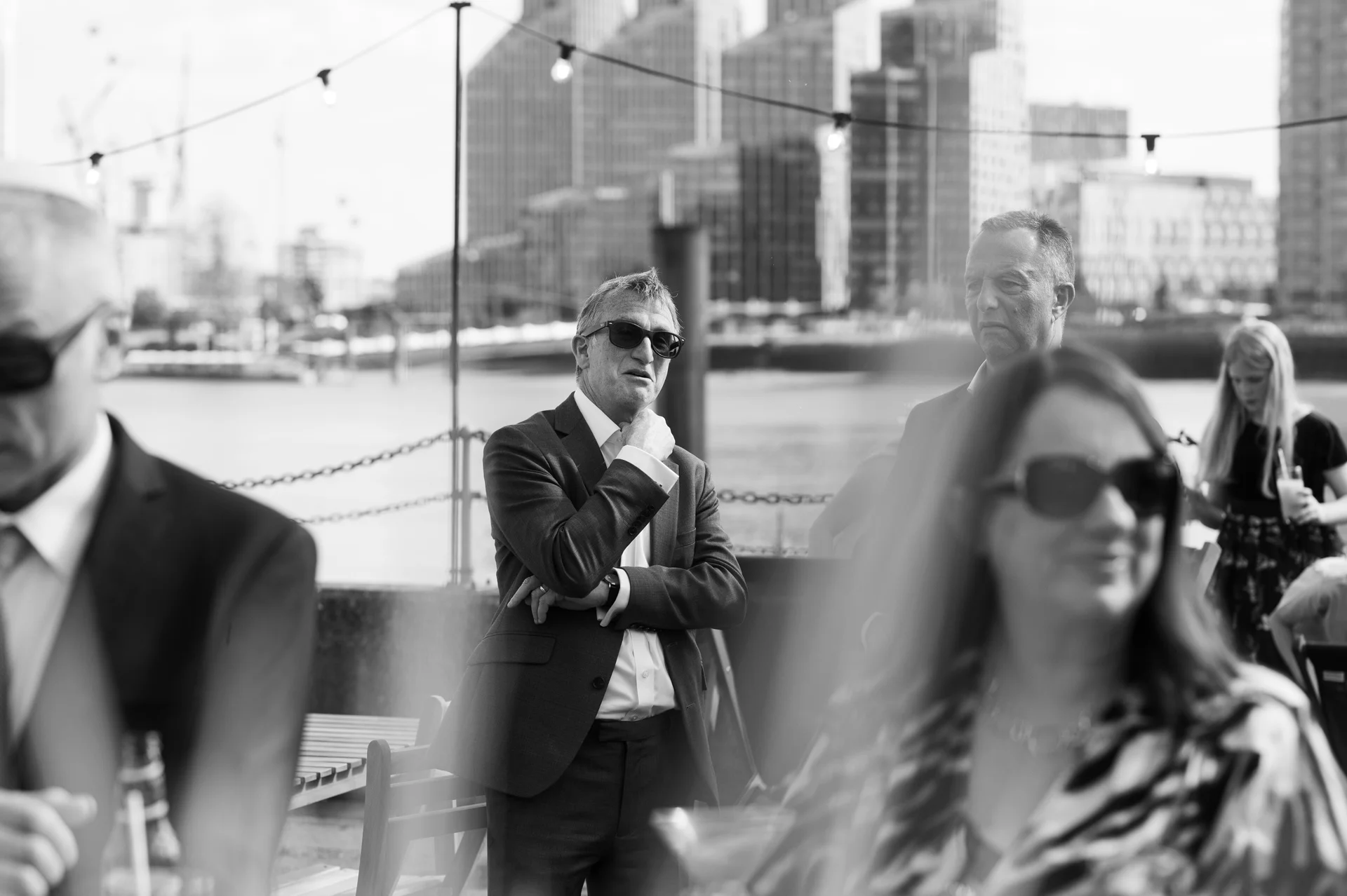 Wedding guests mingle on a city rooftop terrace with a riverside backdrop under strung festoon lighting in this monochrome candid shot.