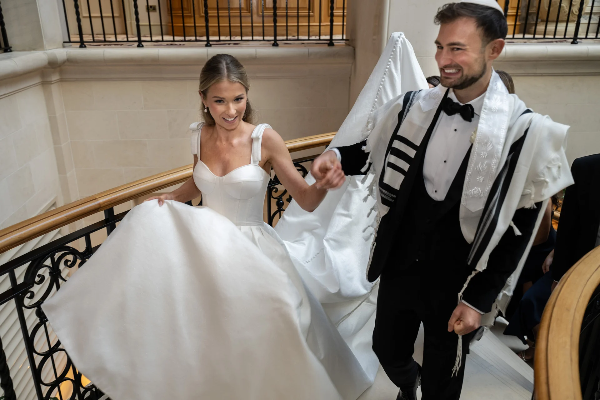 A smiling bride holding her voluminous white gown and a groom wearing a tuxedo and tallit walk down a grand sweeping staircase together.
