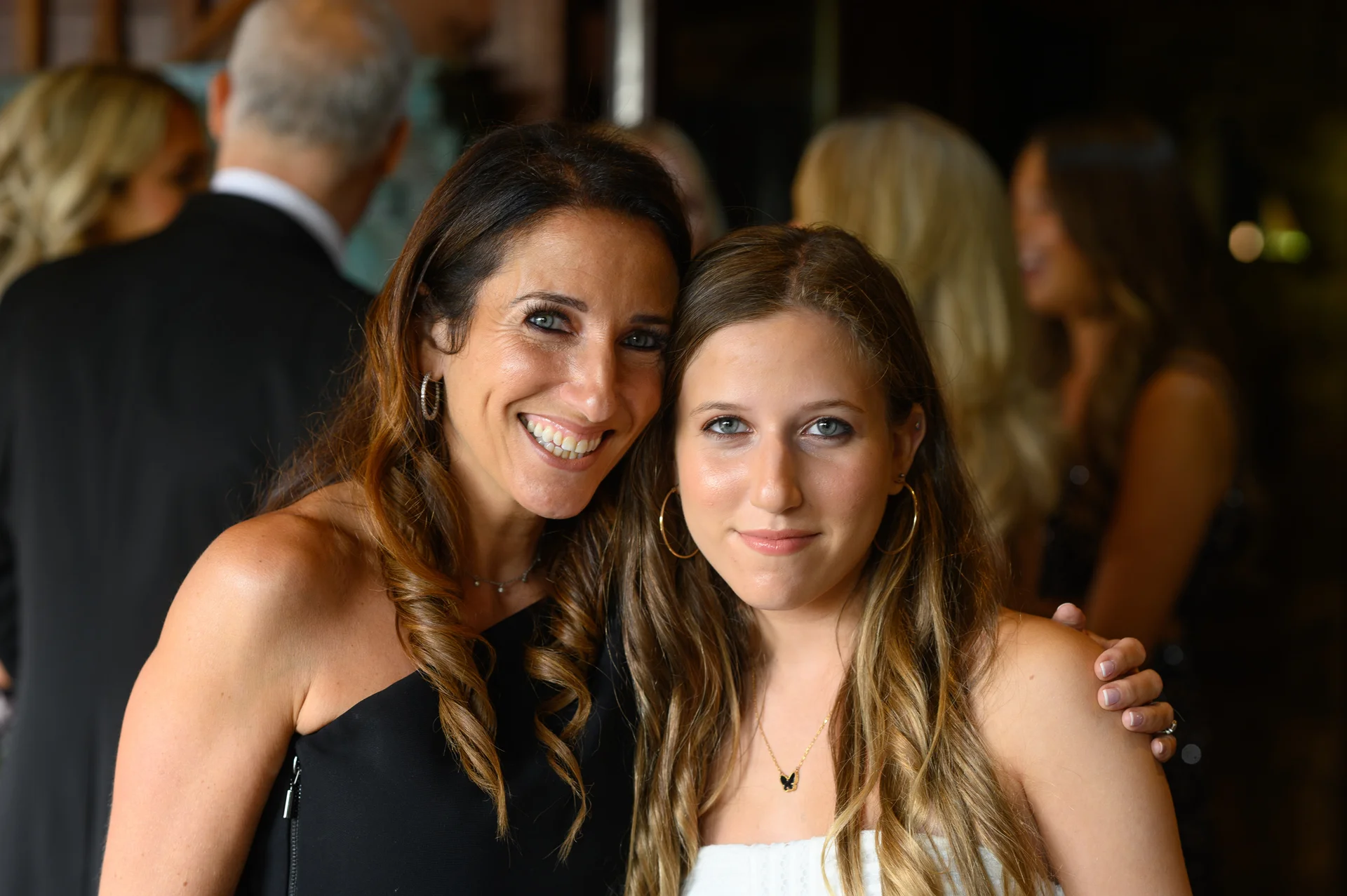 Two female wedding guests pose closely together smiling at the camera during an evening reception.
