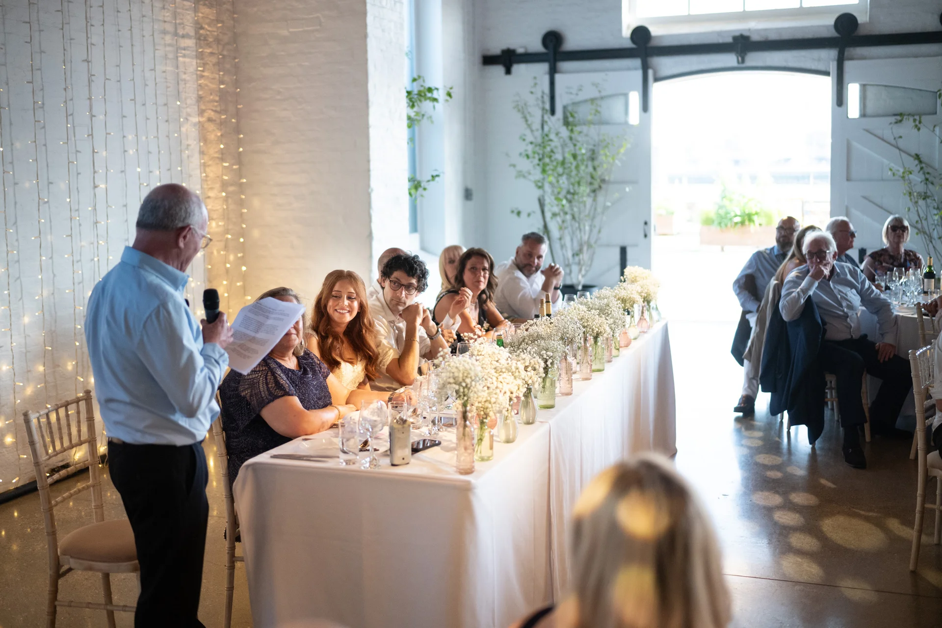 A guest delivers a speech holding notes at a long top table decorated with baby's breath floral arrangements in an industrial warehouse wedding venue.