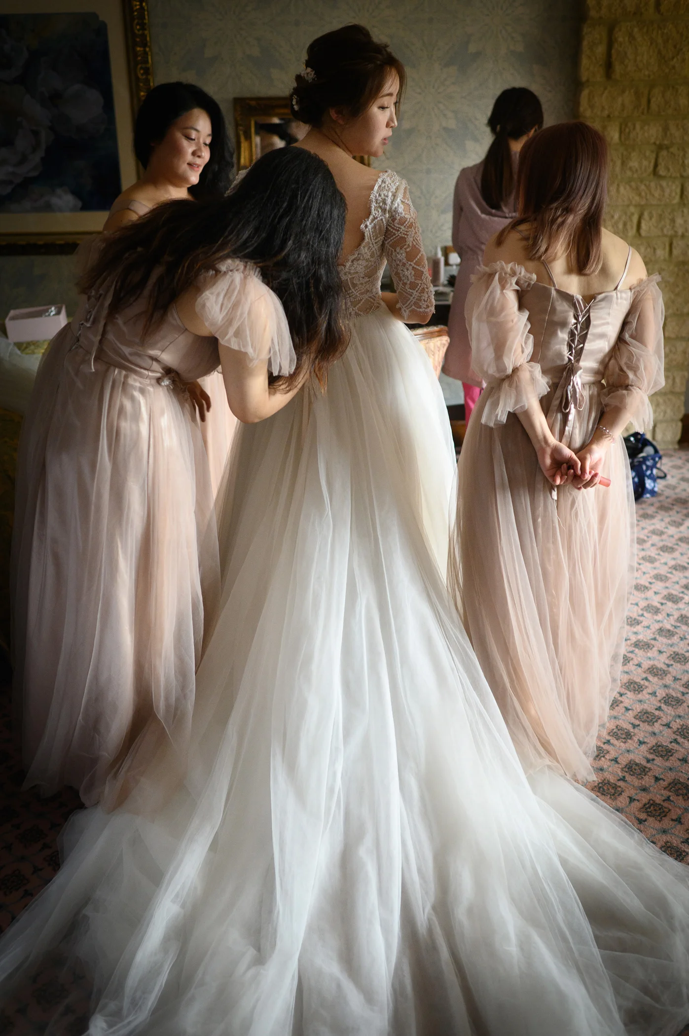 Bridesmaids help the bride button and adjust her lace-detailed wedding dress with a dramatic tulle skirt during morning preparations.
