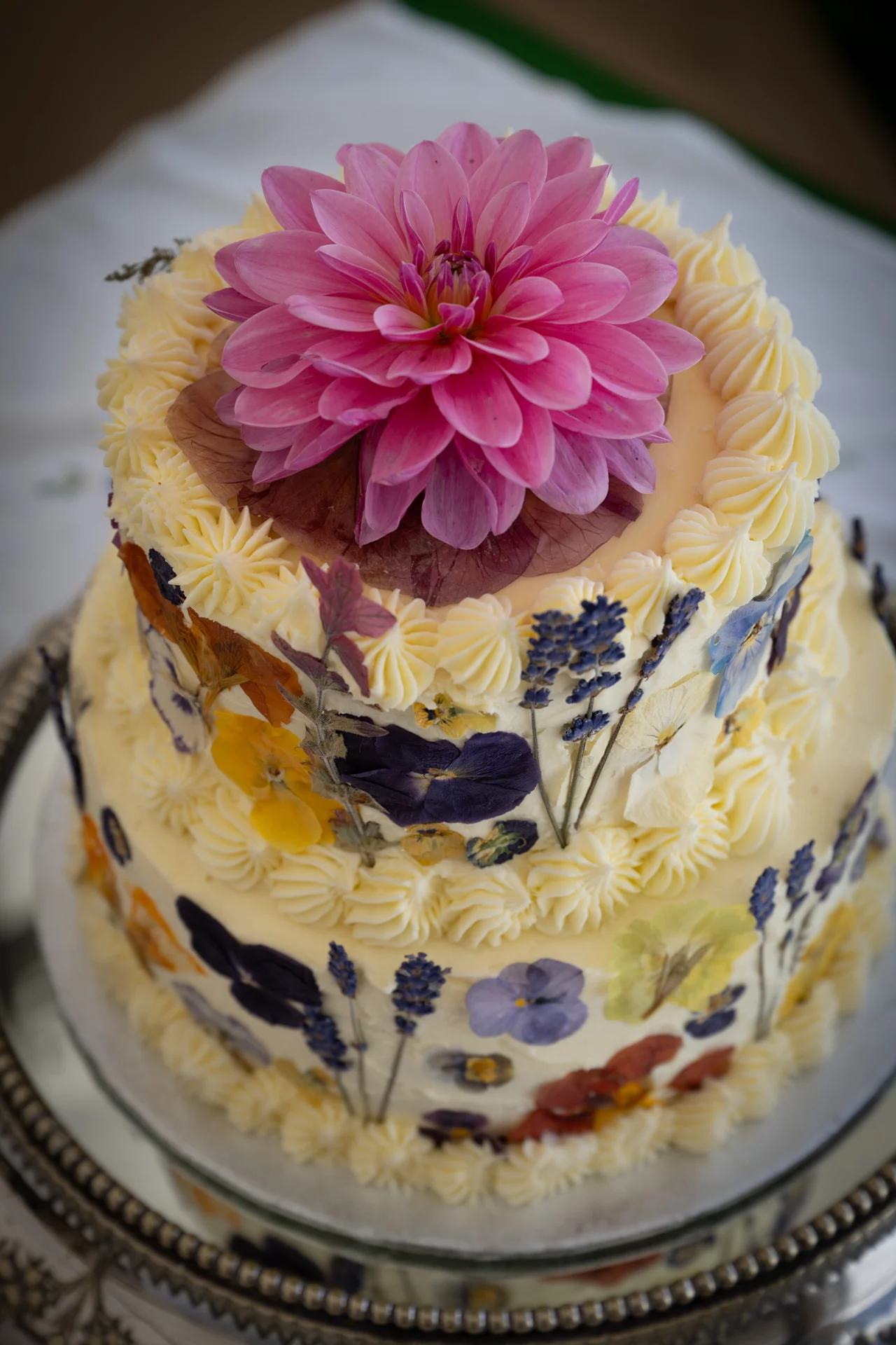A two-tier buttercream wedding cake decorated with pressed flowers along the sides and a large pink dahlia on top, captured in a close-up shot.