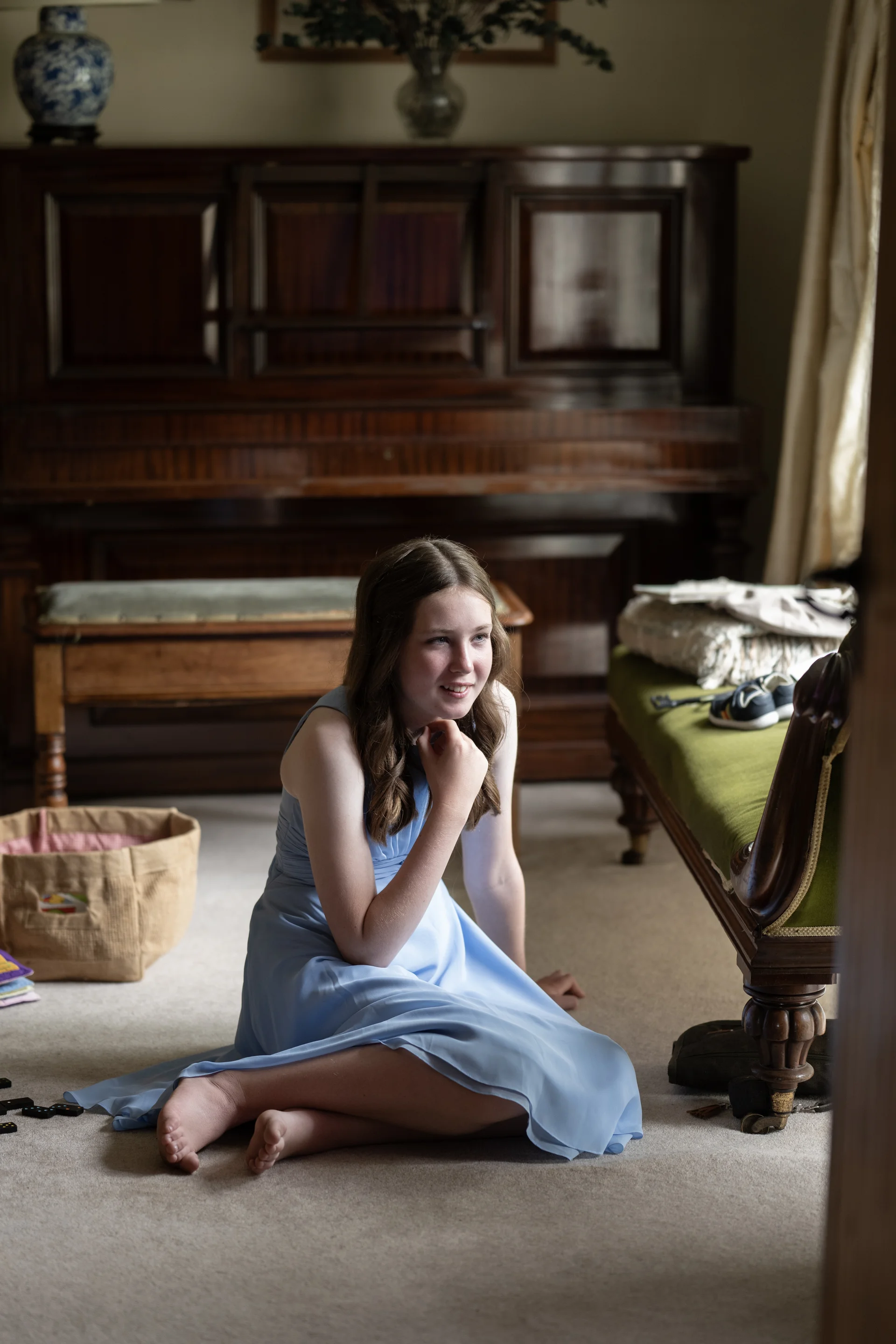 A young bridesmaid wearing a light blue dress sits barefoot on the carpet in a room with a piano during wedding morning preparations.