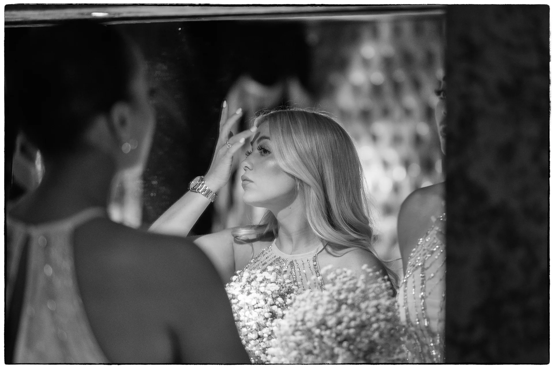 A black and white reflection shot captures a bridesmaid touching her forehead while checking her appearance in a mirror during morning bridal preparations.