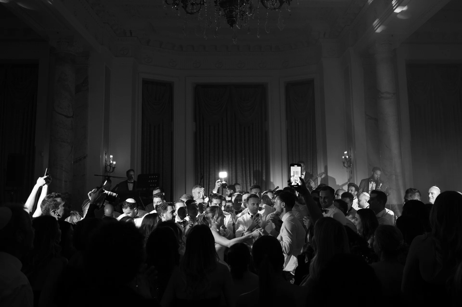 A dramatic black and white shot captures a lively wedding reception in a grand ballroom as guests gather around the couple dancing on a packed dancefloor.