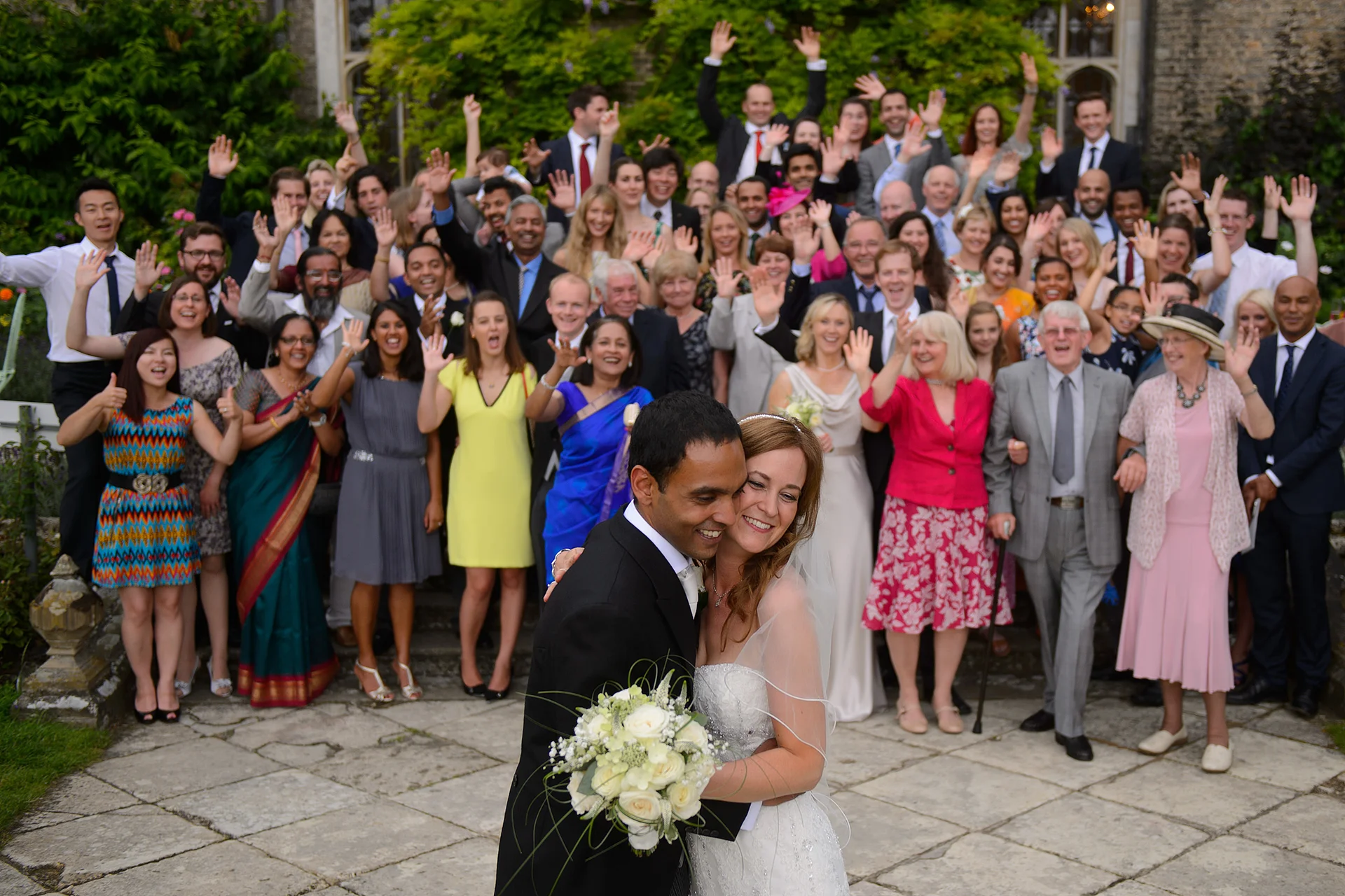 The newly married couple embrace in the foreground while their cheering wedding guests wave and celebrate on stone steps in the background.