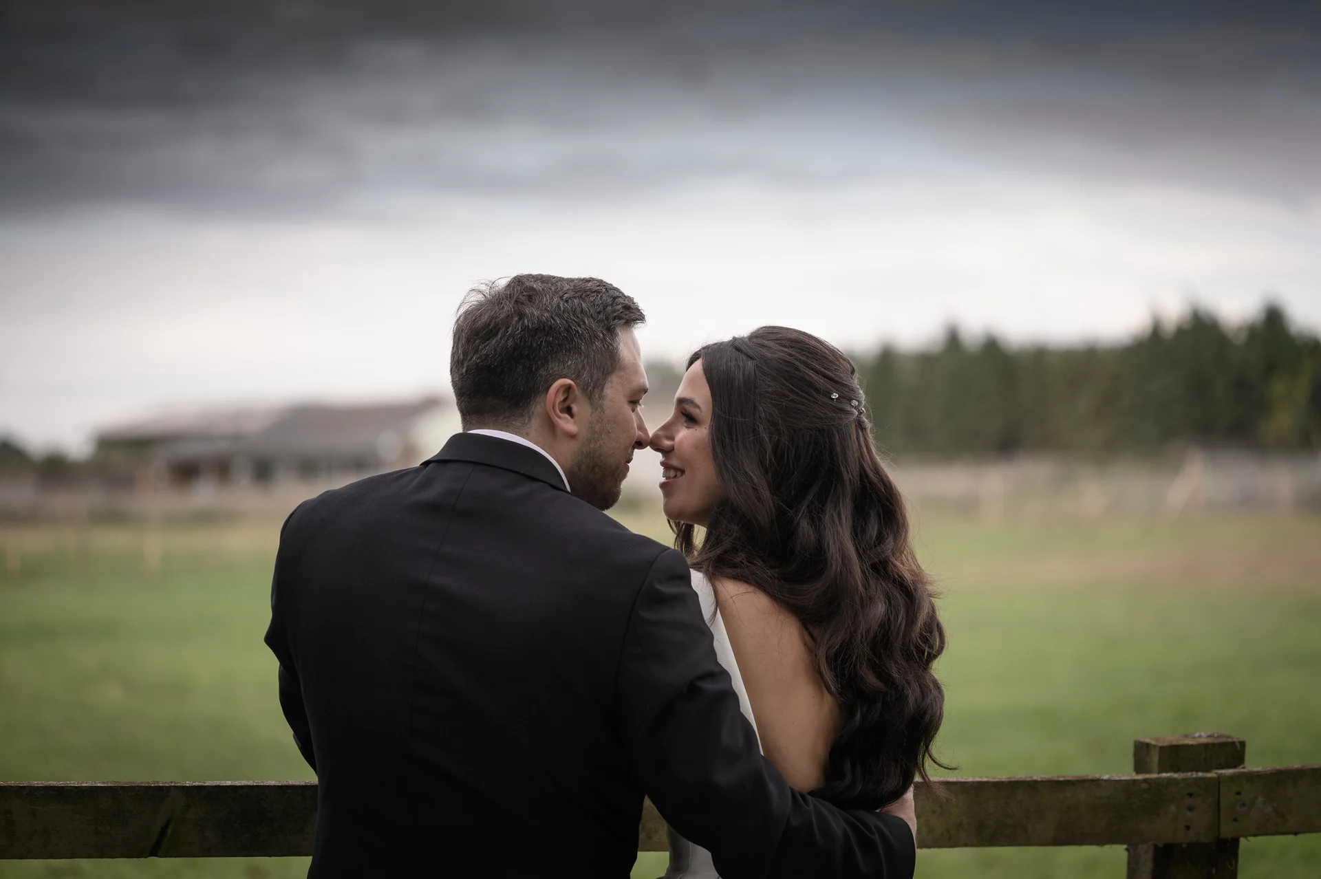 A bride and groom share a tender nose-to-nose moment leaning against a wooden fence with an overcast rural landscape in the background.